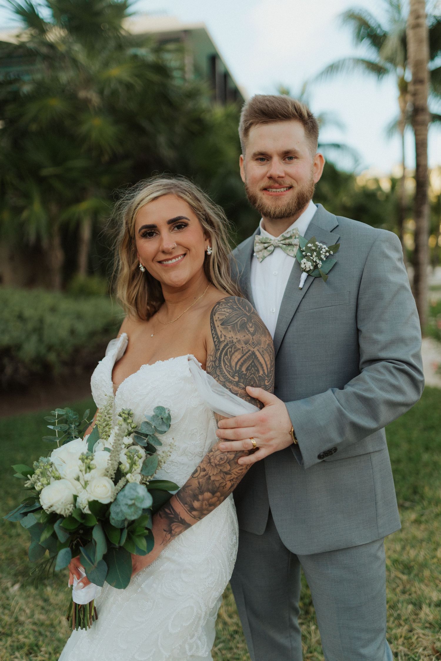 Smiling bride with tattoo sleeve holding white bouquet posing with groom in gray suit outdoors.