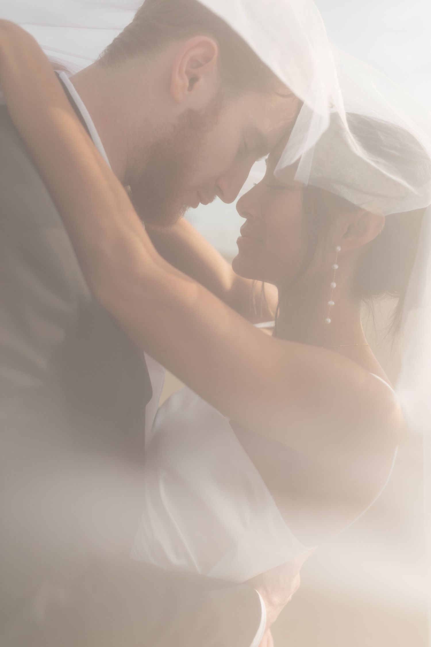 Intimate close-up of bride and groom touching foreheads under flowing wedding veil in soft dreamy light.