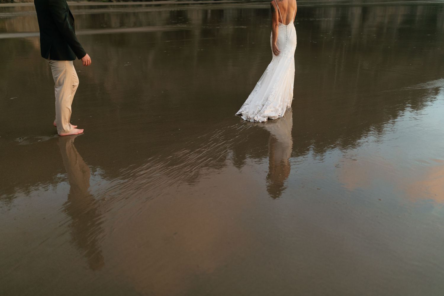 Bride in white gown and groom walk barefoot on wet reflective sand at the beach.