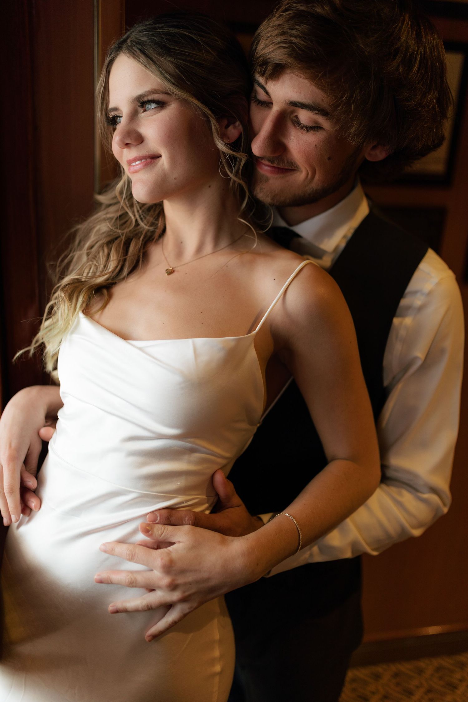 Groom tenderly embracing bride from behind in warm indoor lighting on their wedding day.