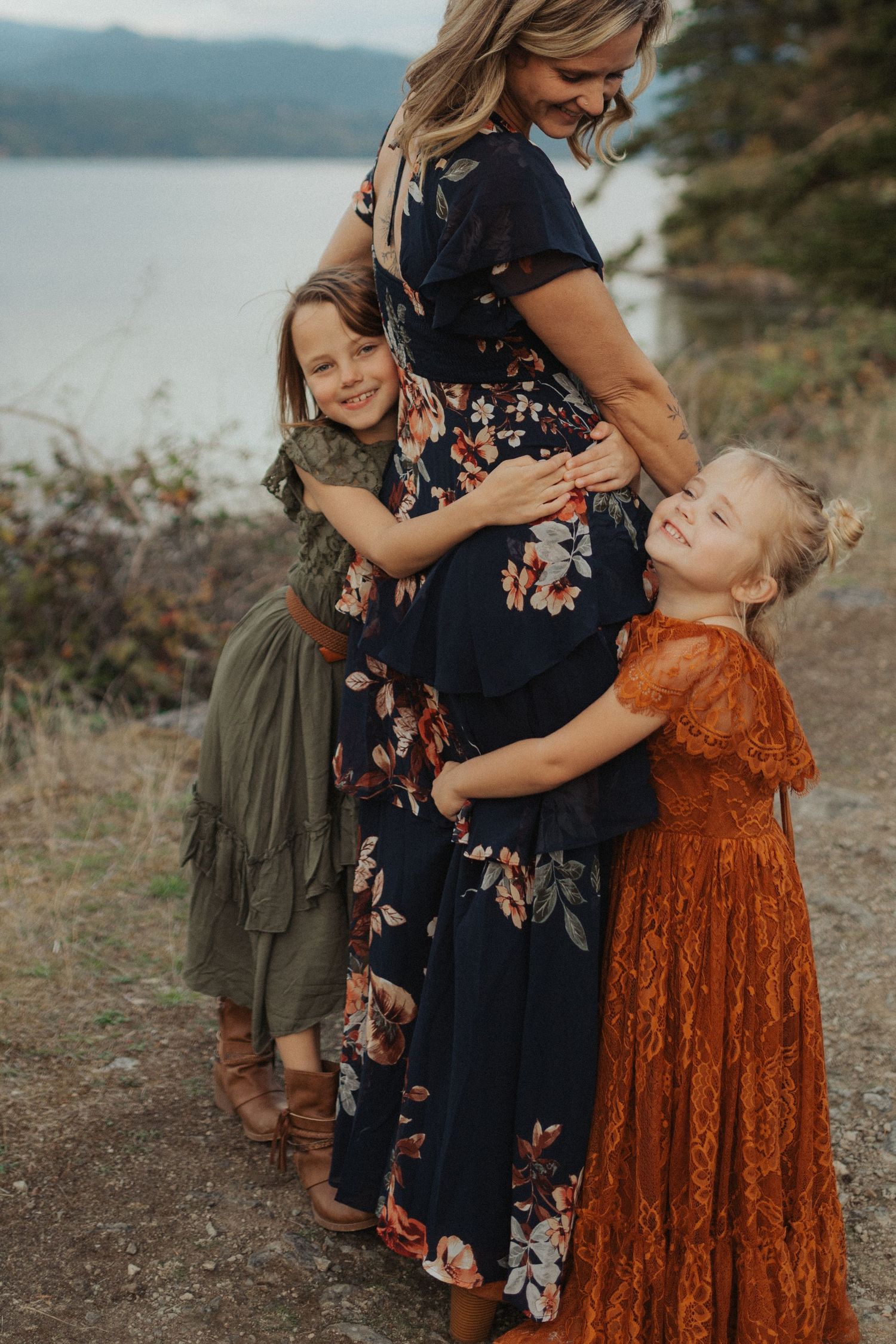 Two young girls lovingly hug their pregnant mothers baby bump outdoors near a mountain lake.