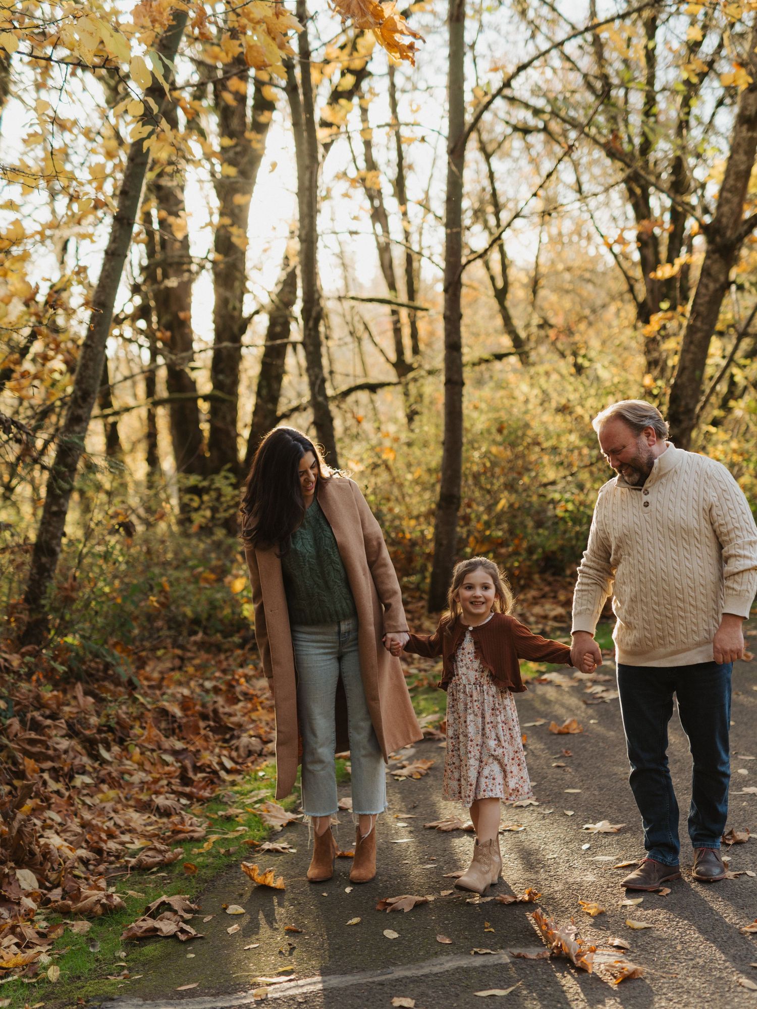 Family of three holding hands on a sunlit autumn path surrounded by golden fall foliage.