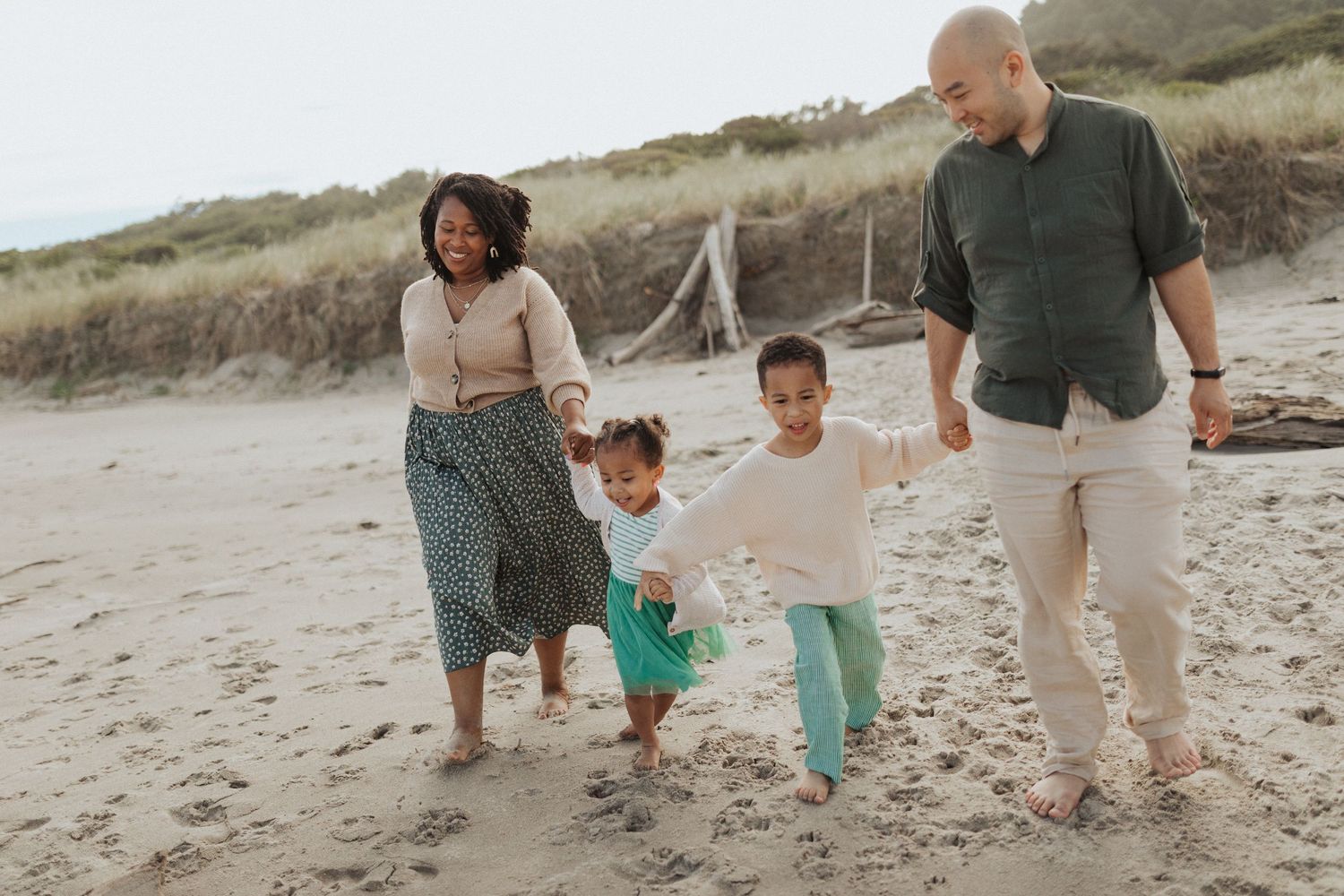 Joyful interracial family of four walking hand-in-hand along a sandy beach with dune grass in background.