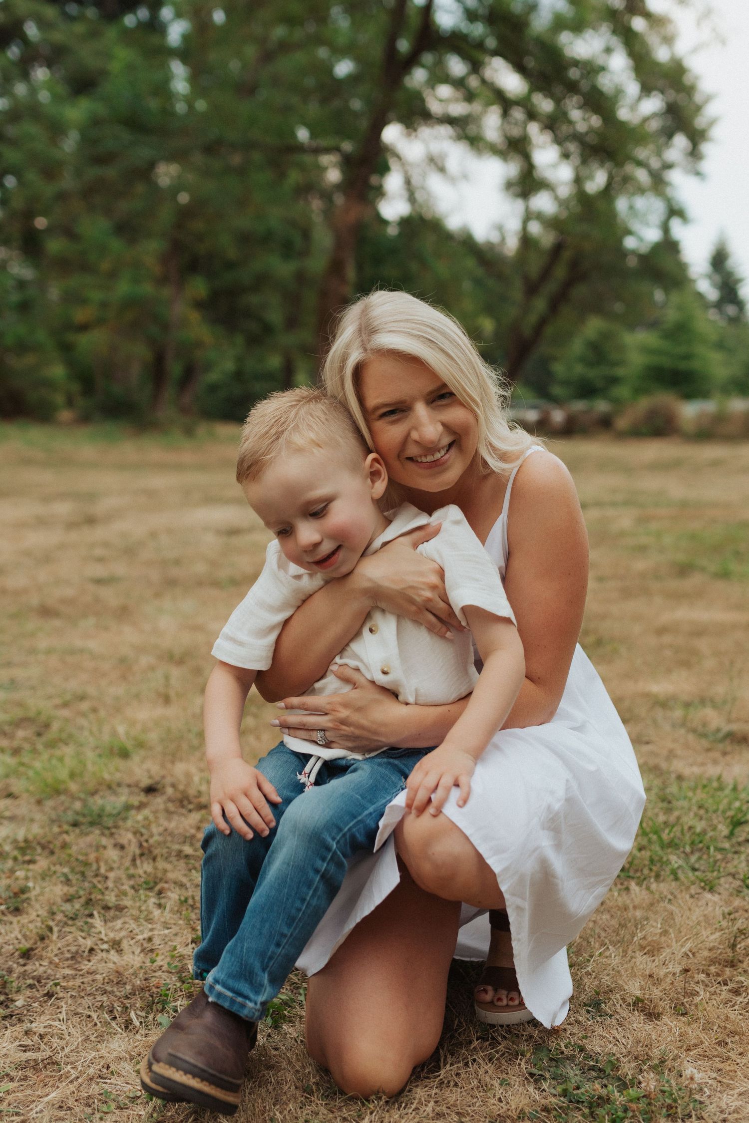 Smiling blonde mother kneeling and hugging her young son outdoors in a grassy park setting.