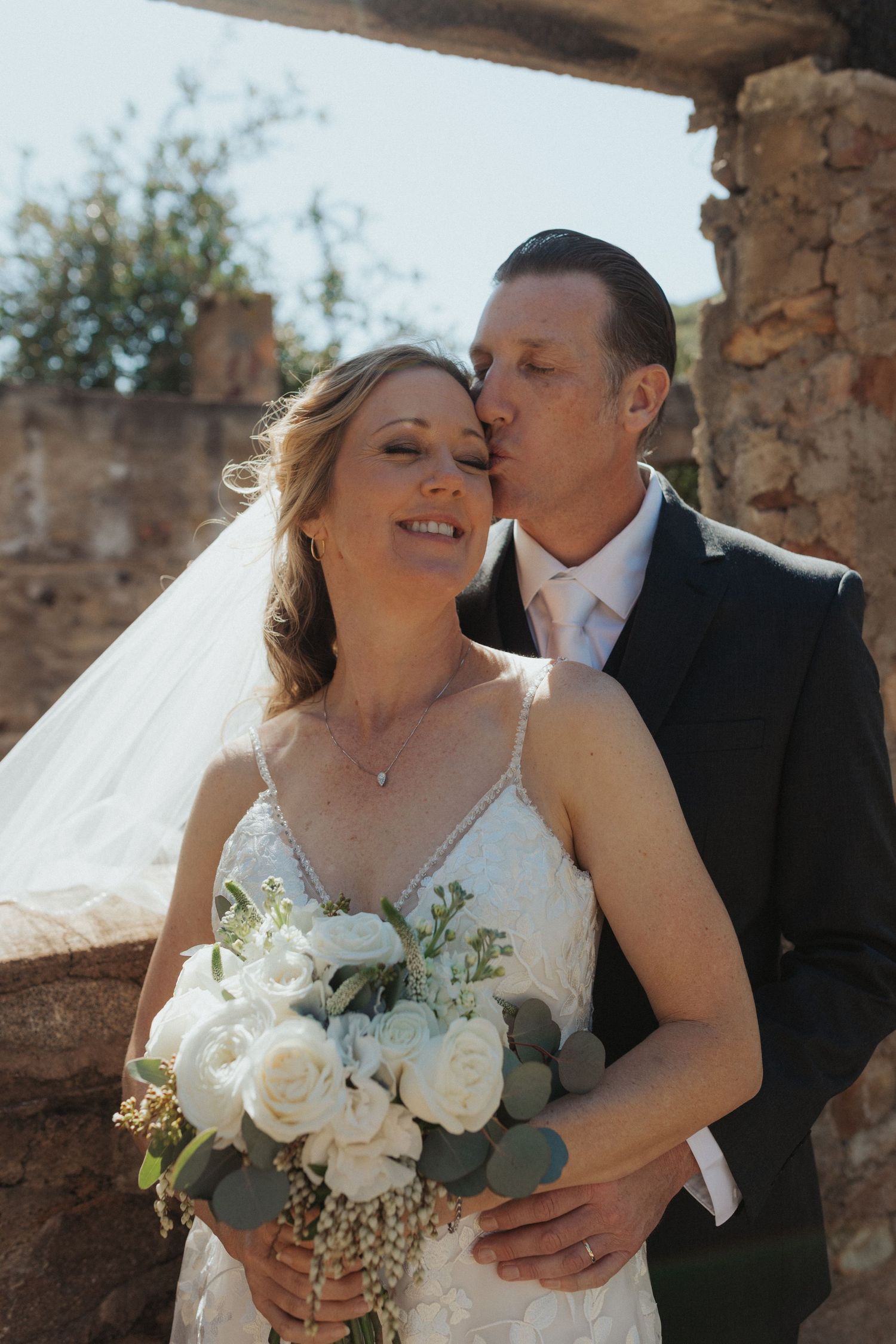 Groom kissing bride on the cheek as she smiles holding a white rose bouquet at a rustic outdoor wedding.