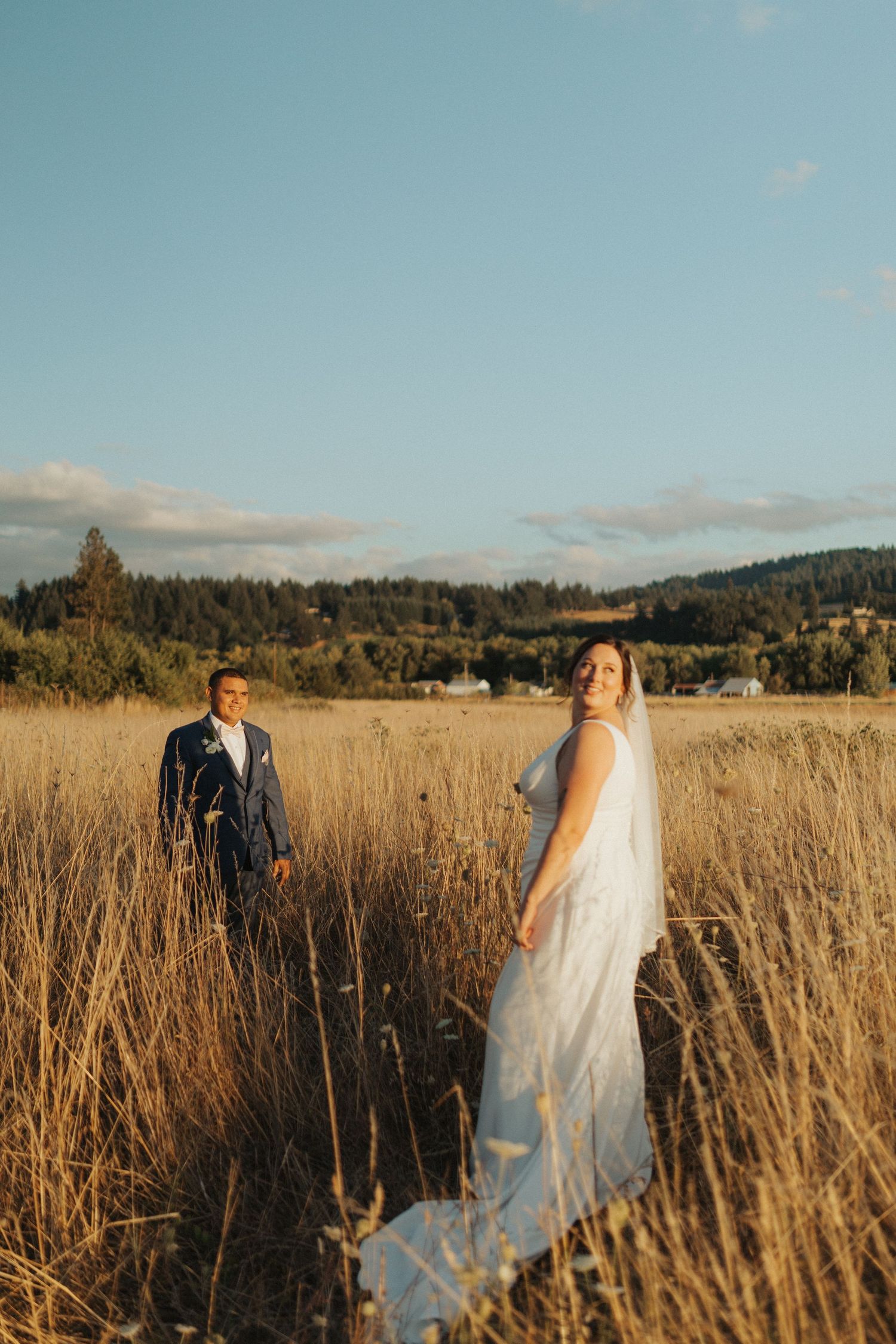 Bride in white gown with veil and groom in blue suit pose in a golden wheat field at sunset.