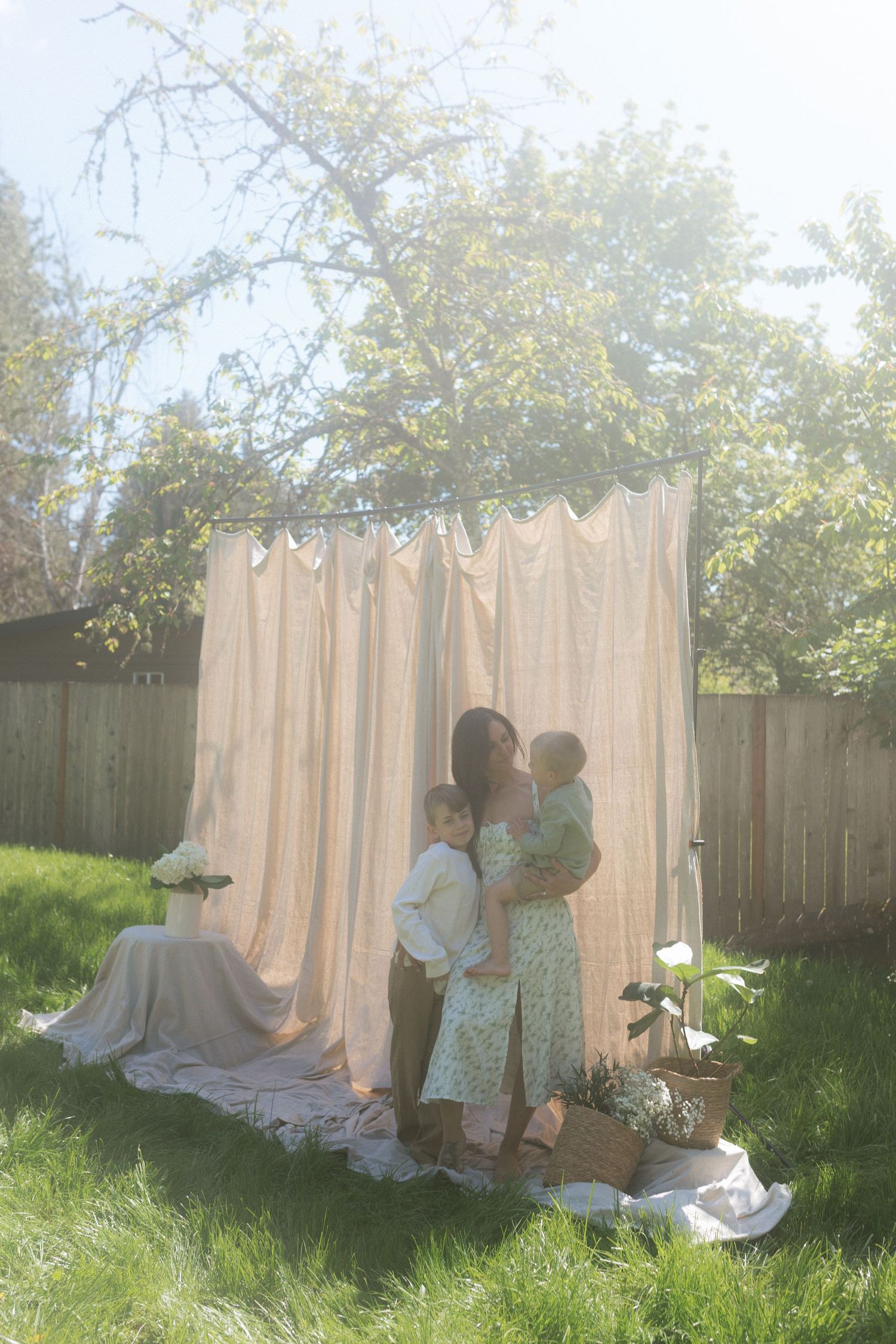 A mother holds her children in a sunlit backyard with a flowing curtain backdrop.