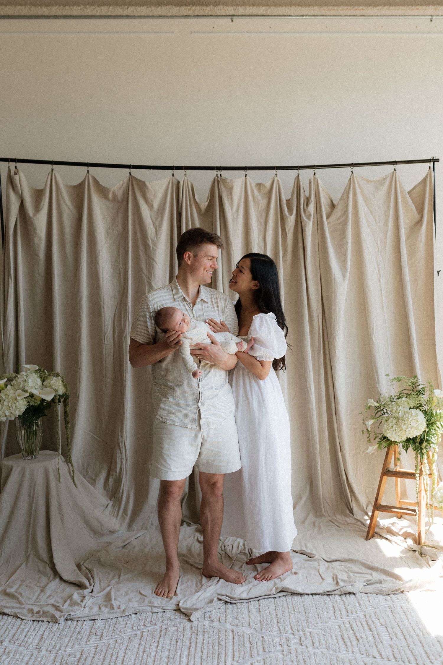 Happy couple in white and beige outfits holding newborn baby in bright studio with floral arrangements.