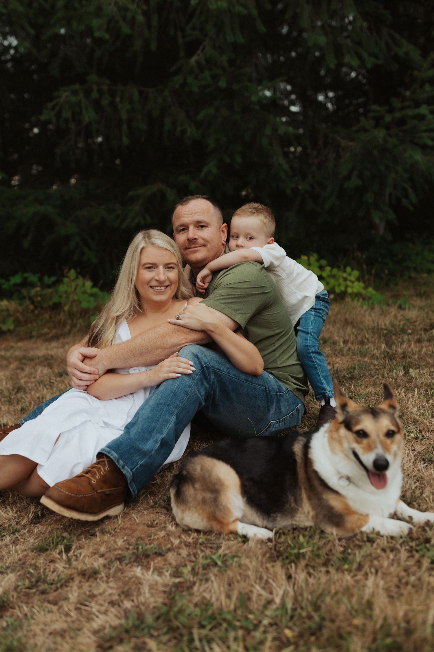 Smiling family with young child and corgi dog relaxing together on dry grass in a park.
