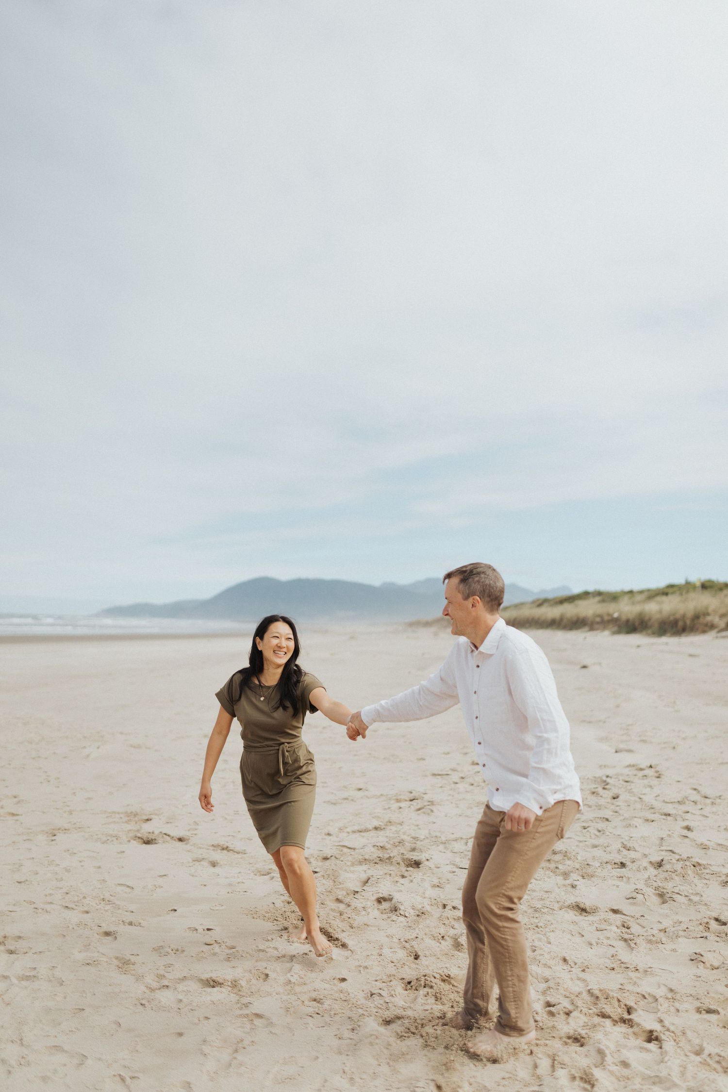 Couple holding hands and walking barefoot on a wide sandy beach on a cloudy day.