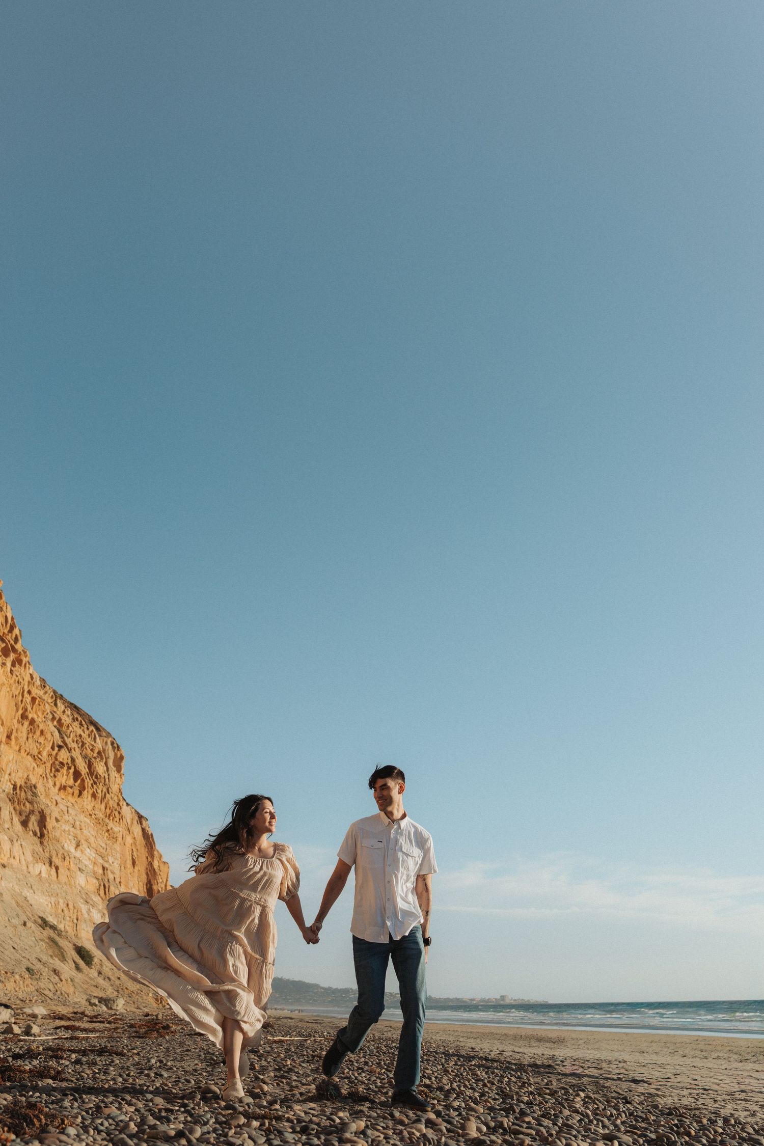 Couple holding hands and walking on rocky beach with towering sandy cliffs under a clear blue sky.