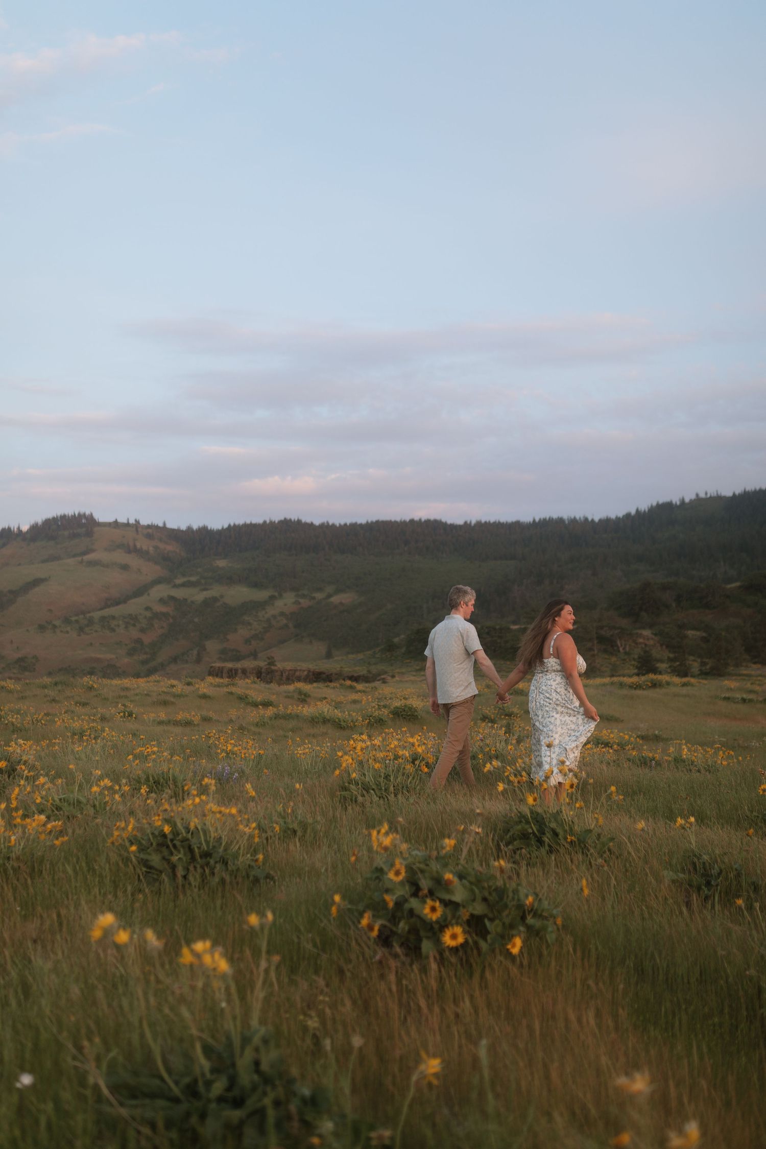 Couple holding hands while walking through a vast meadow of golden sunflowers beneath a soft evening sky.