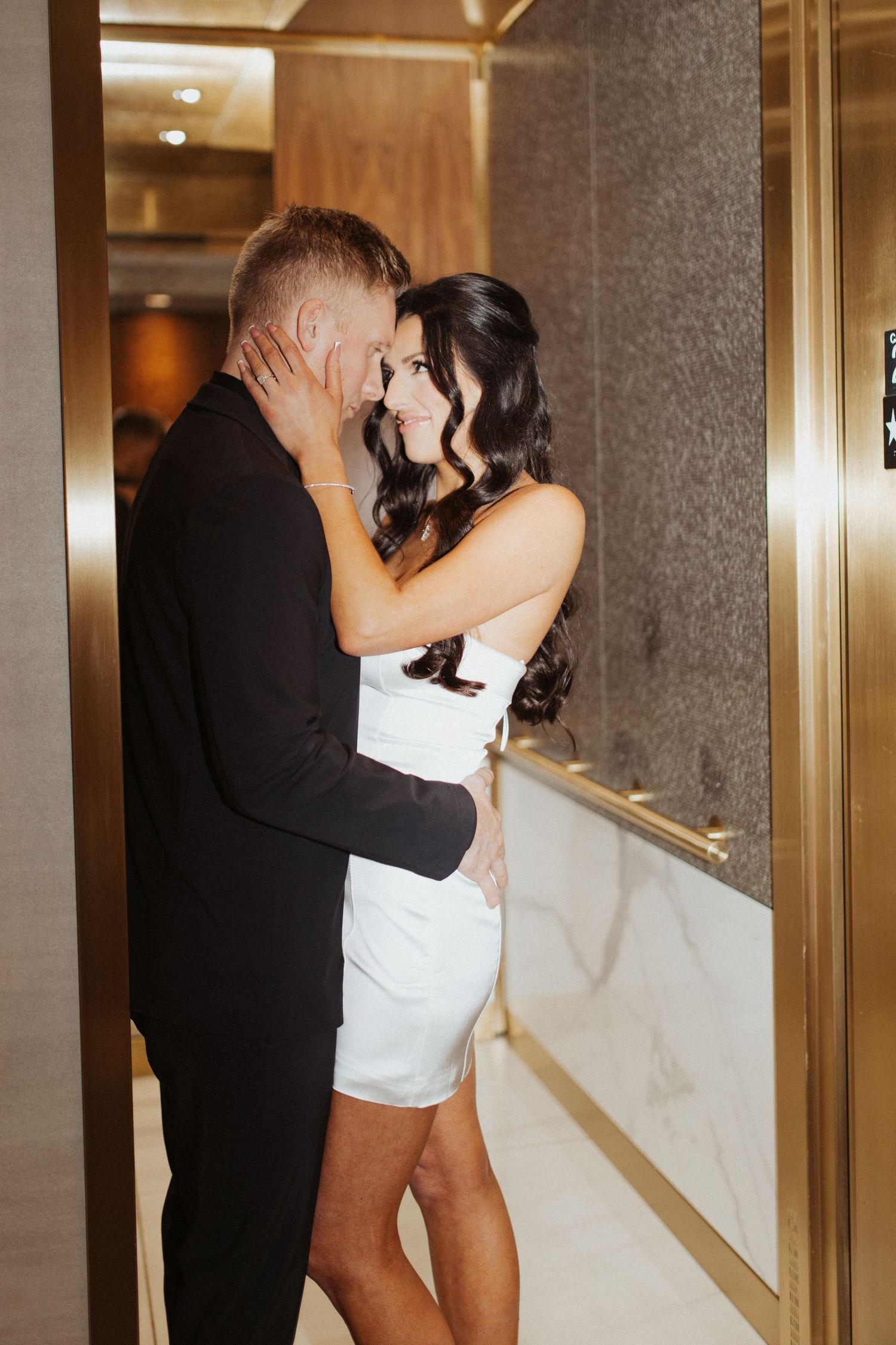 Couple in elegant attire sharing a romantic moment in a hotel elevator, woman in white dress, man in black suit.