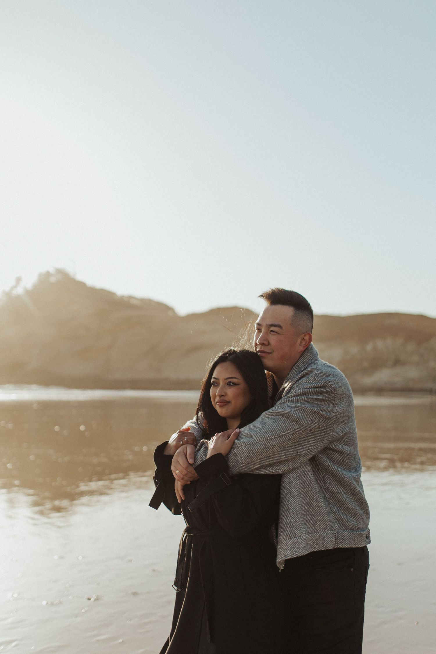 Couple embracing on sunlit beach with sandy dunes and clear sky during engagement photo session.