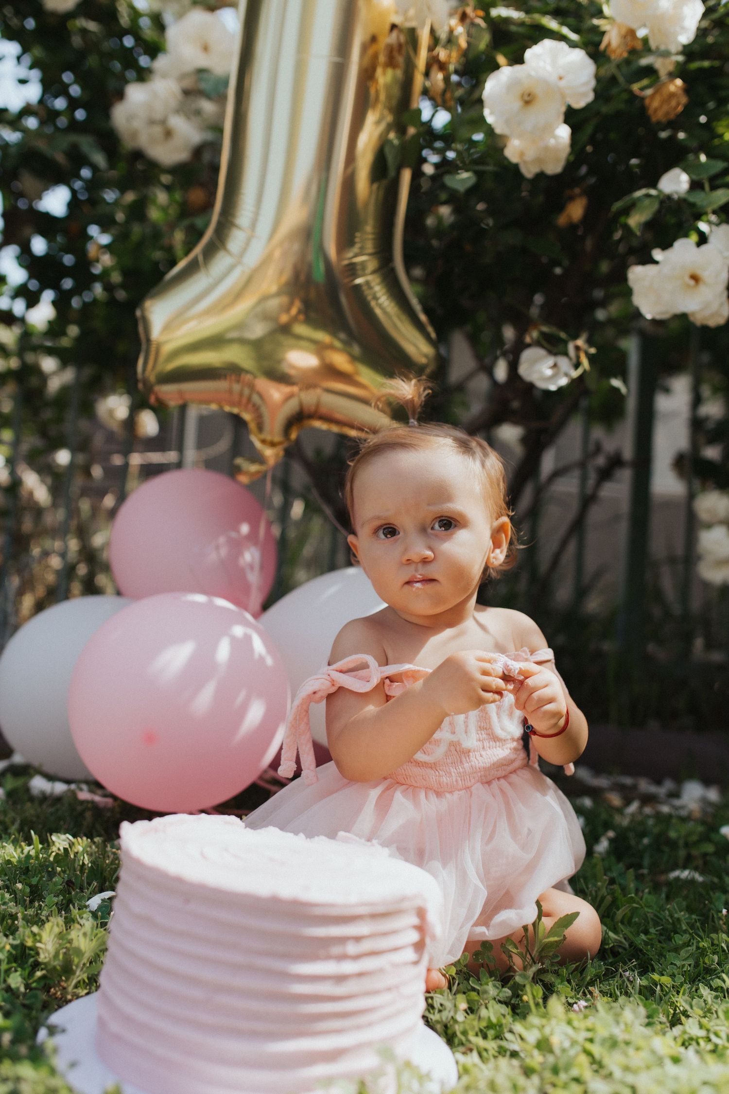 One-year-old in pink dress sits near smash cake with gold number one balloon and pink balloons.
