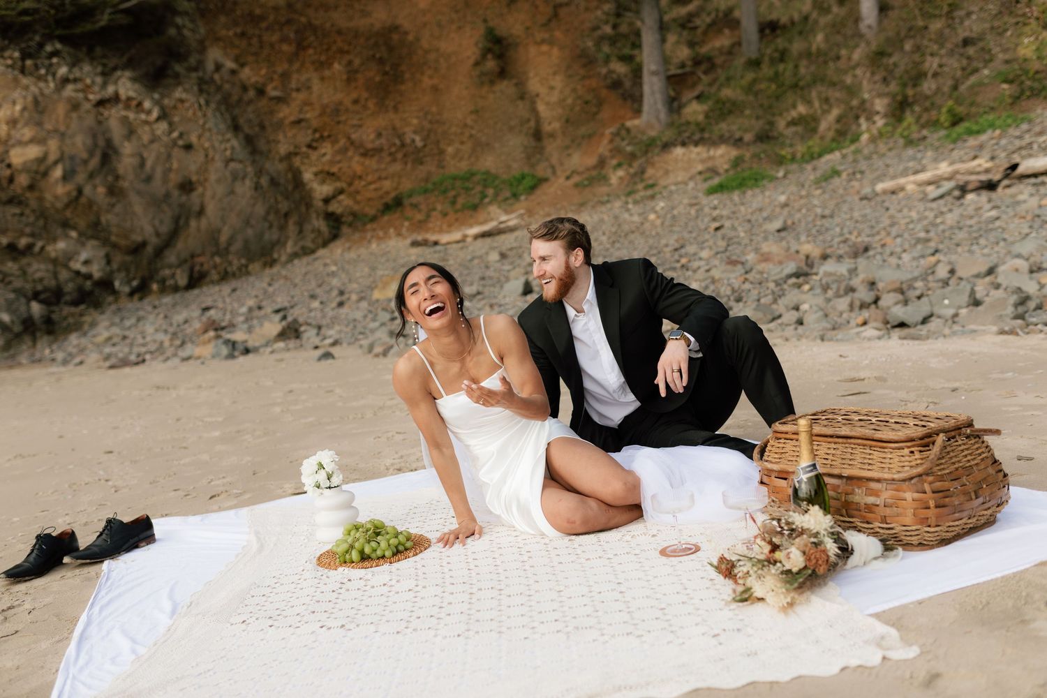 Laughing bride and groom enjoy a beach picnic with champagne and a wicker basket on a white blanket.