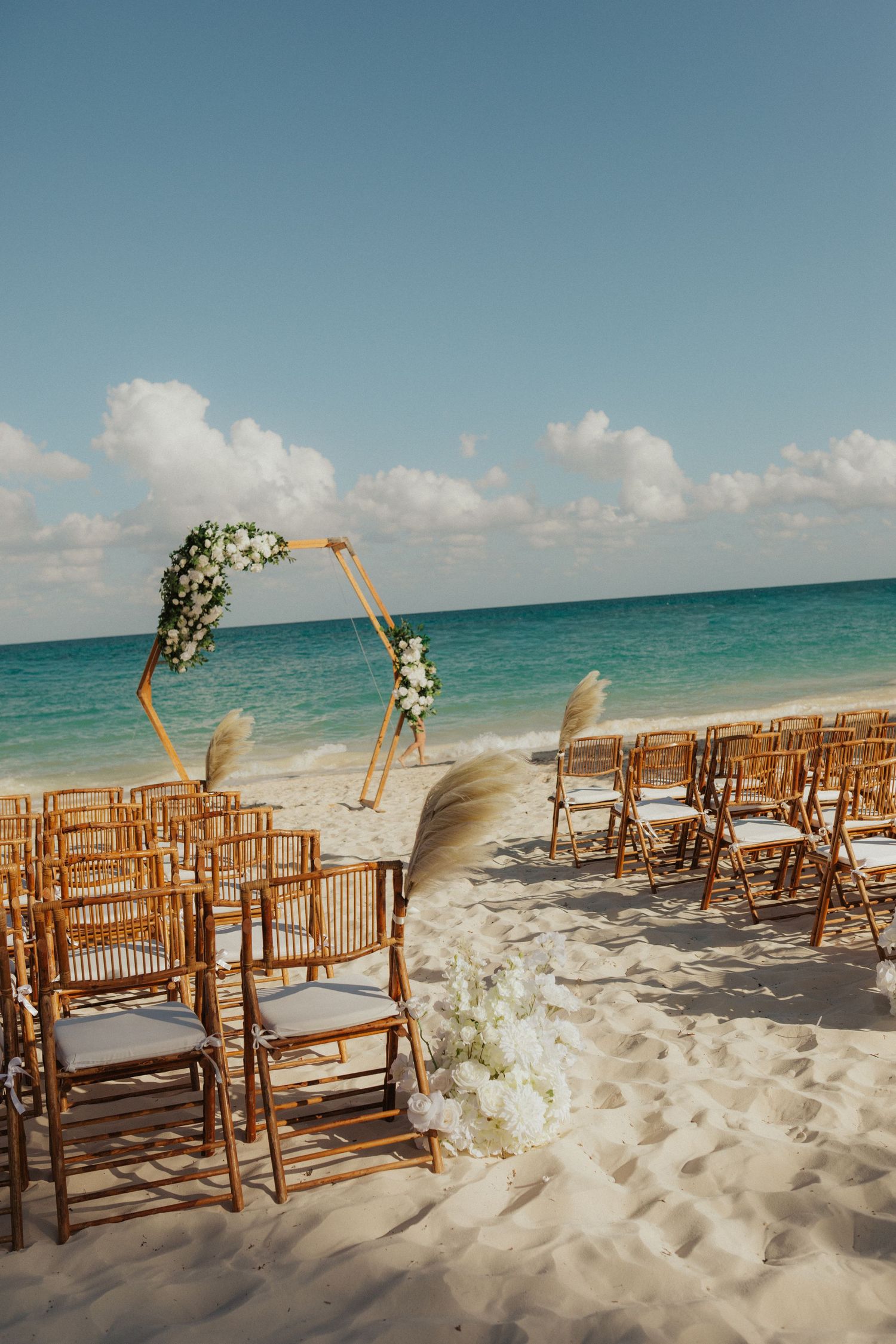 Beach wedding ceremony setup with bamboo chairs, floral arch, and turquoise ocean backdrop.