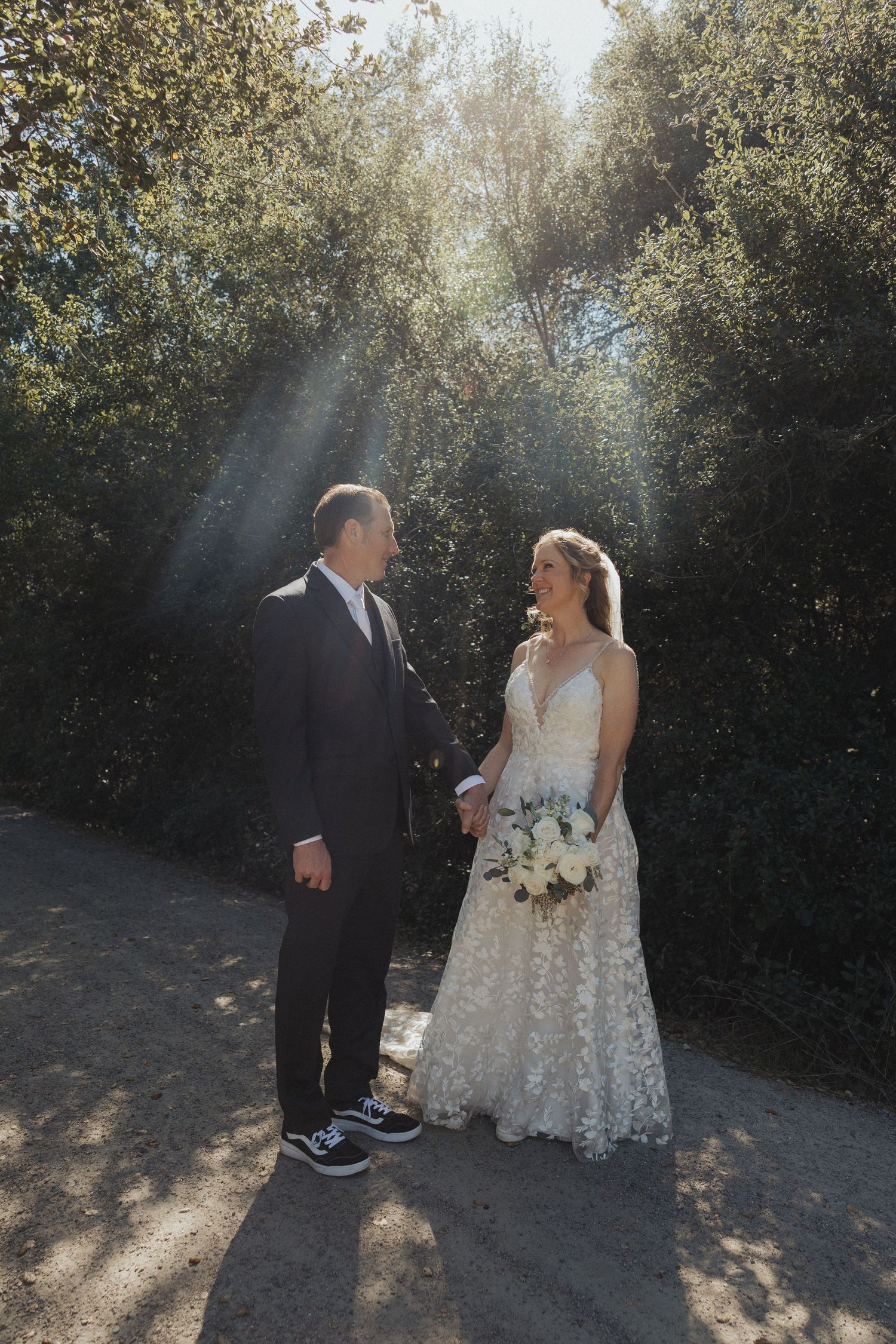 Bride and groom smile at each other holding hands on a gravel path with sunbeams behind them.