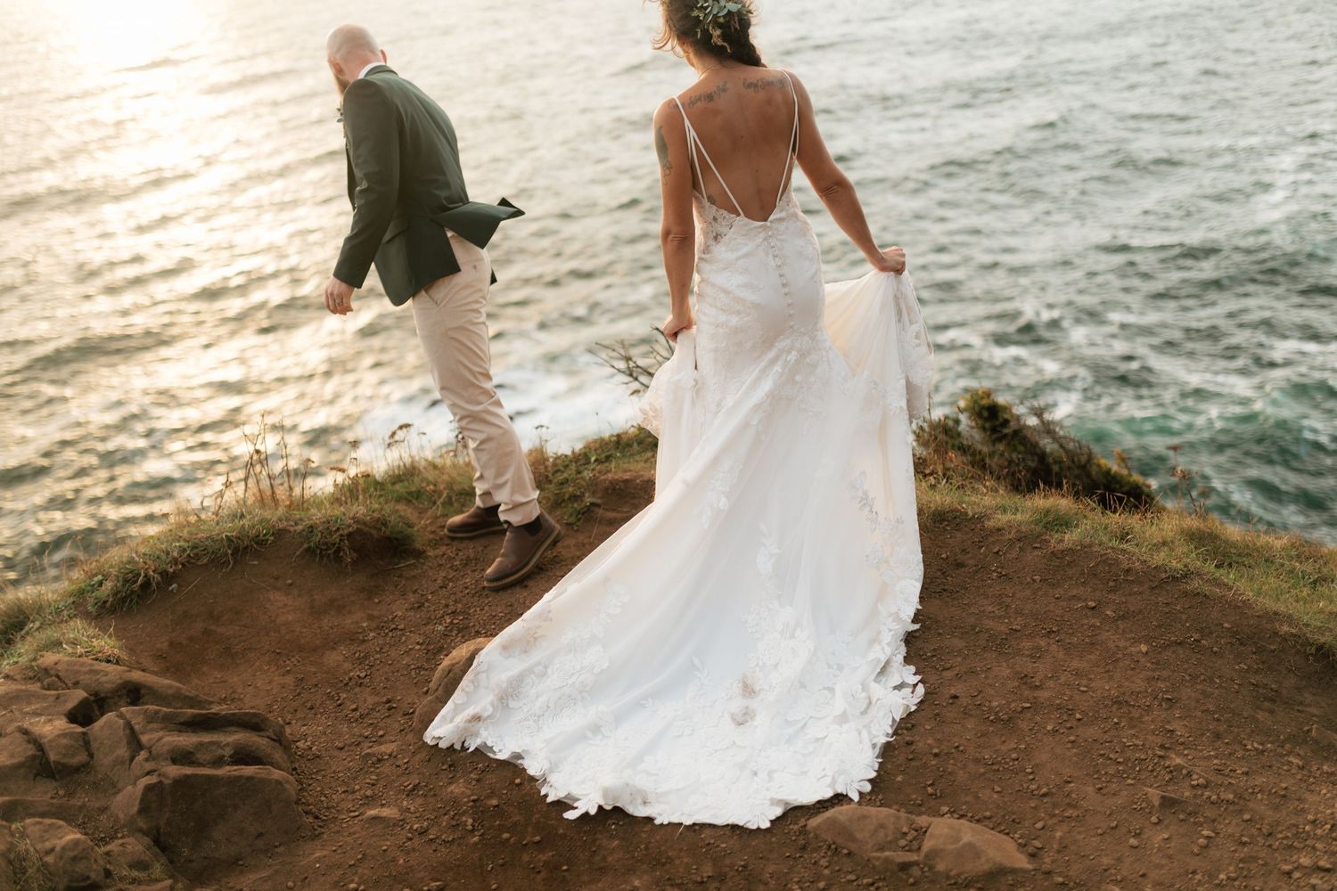 Bride in lace gown with long train standing on a cliffside overlooking the sea.