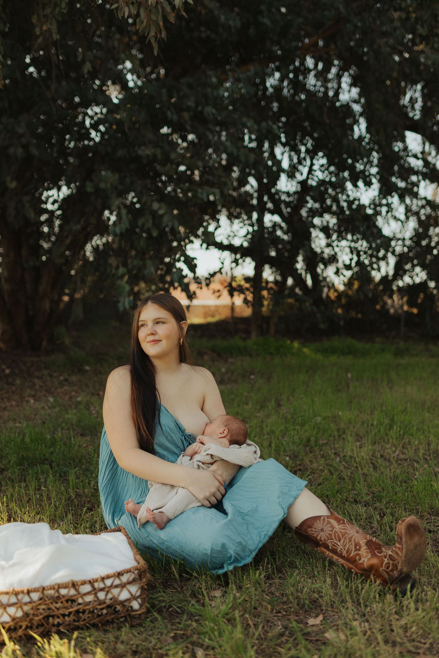 Young mother in teal dress nursing newborn baby outdoors on green grass with wicker basket nearby.