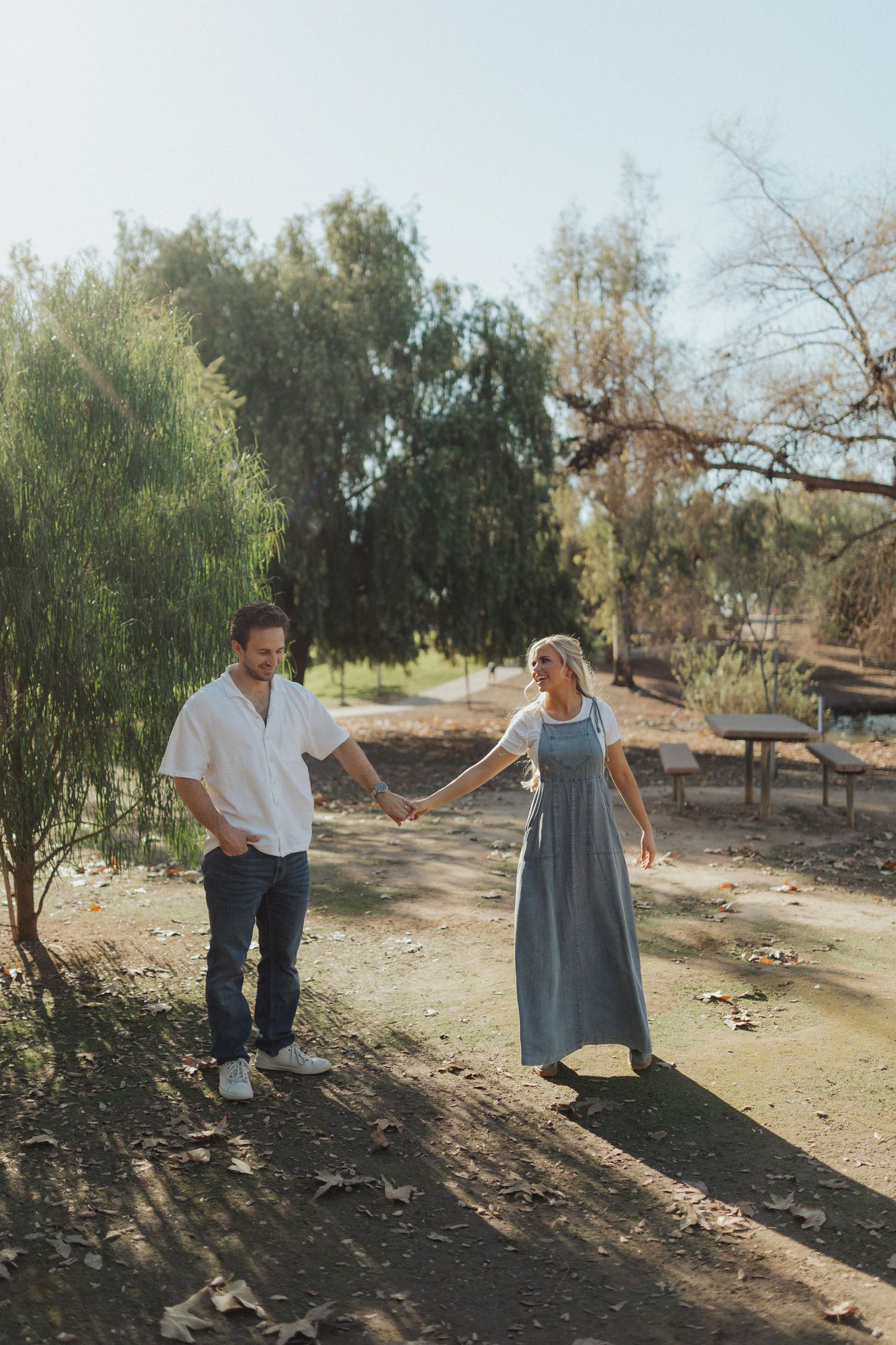 A couple holds hands and walks together along a sunlit park path surrounded by green trees.