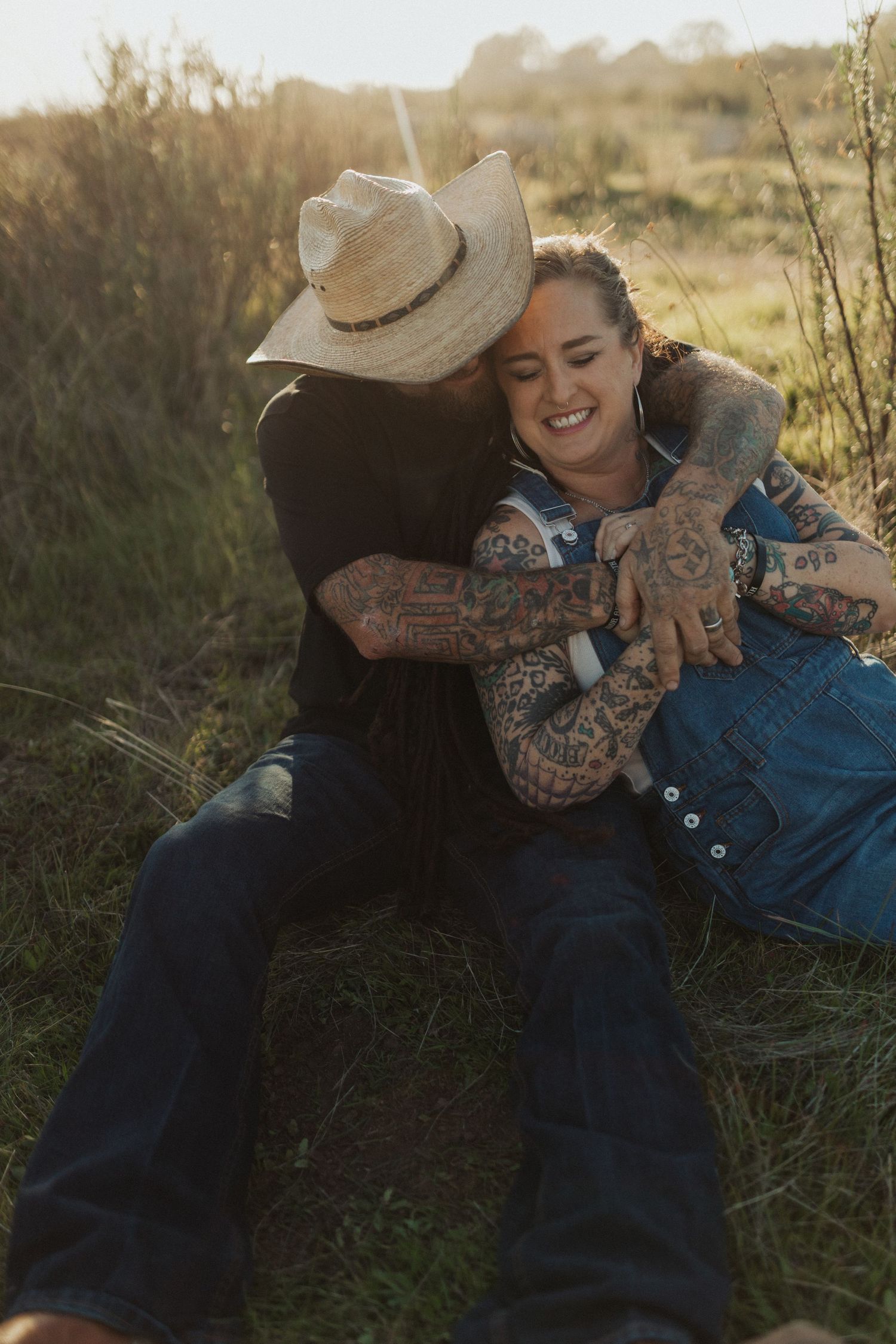 Tattooed couple sitting in golden grass field, man in cowboy hat embracing smiling woman at sunset.