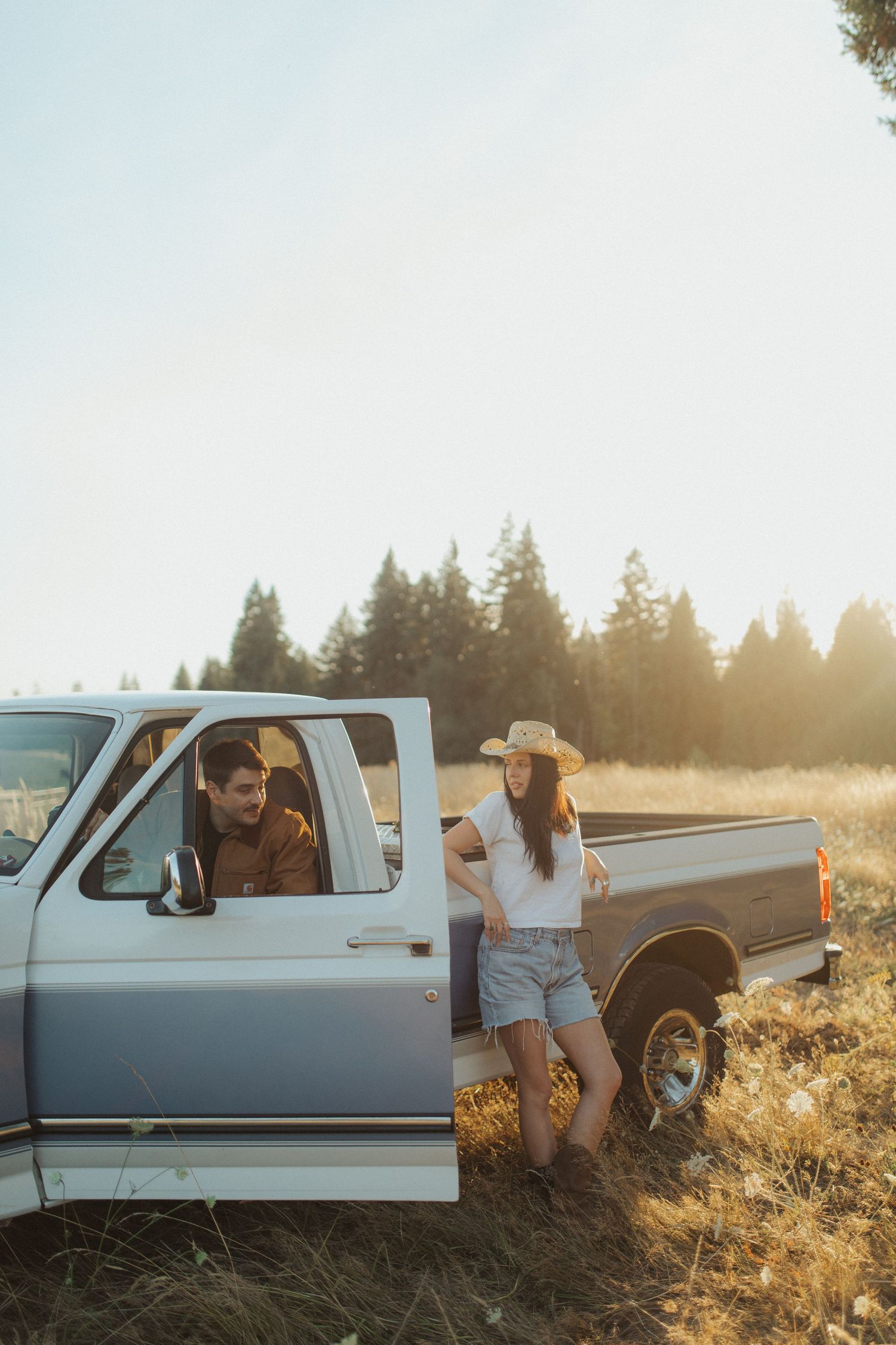 A couple shares a relaxed moment by a vintage blue pickup truck in a sunlit meadow surrounded by pine trees.
