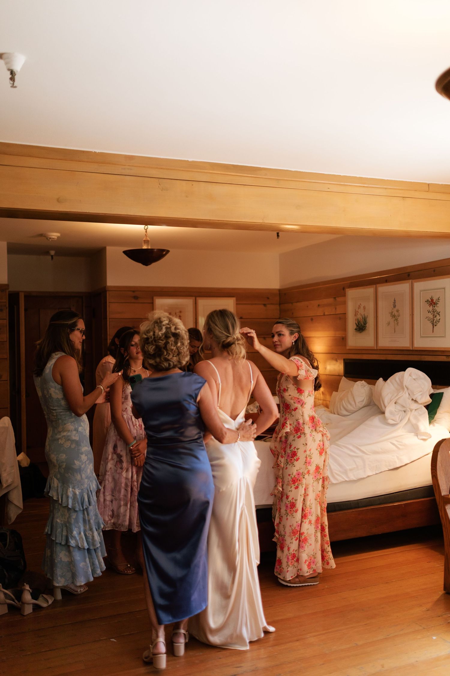 Bridesmaids and family help bride into her white wedding gown in a warmly lit wood-paneled hotel room.