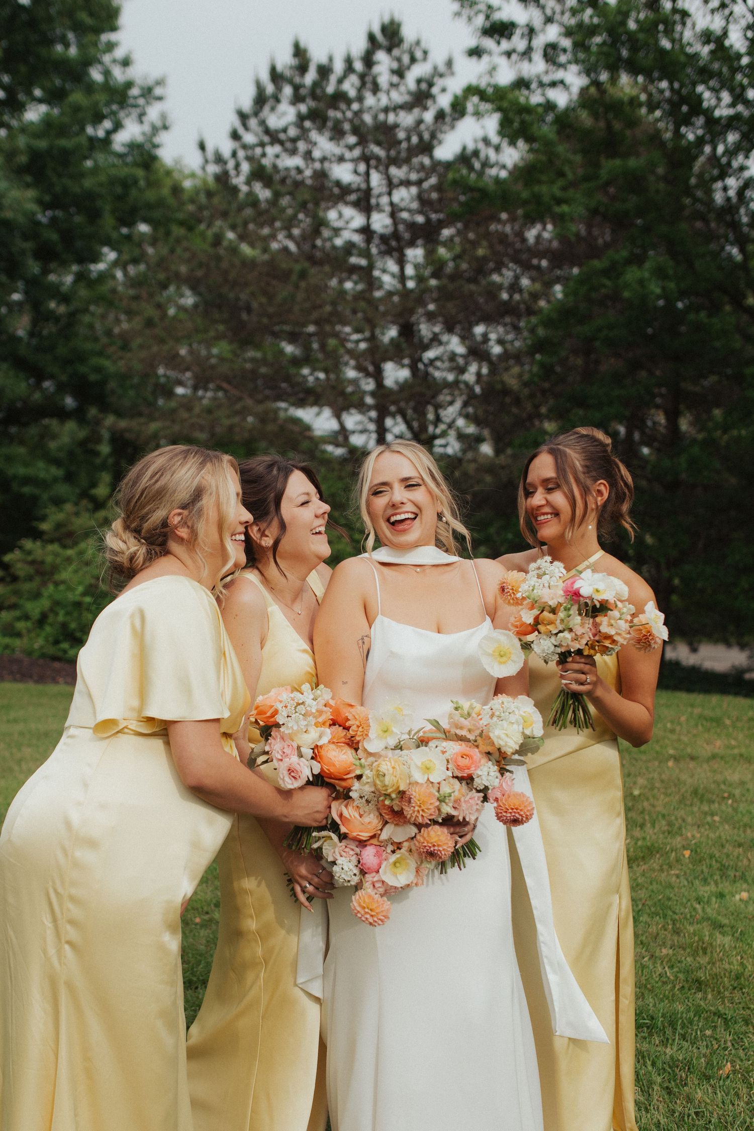 Bride laughs joyfully with three bridesmaids in yellow dresses holding colorful floral bouquets.