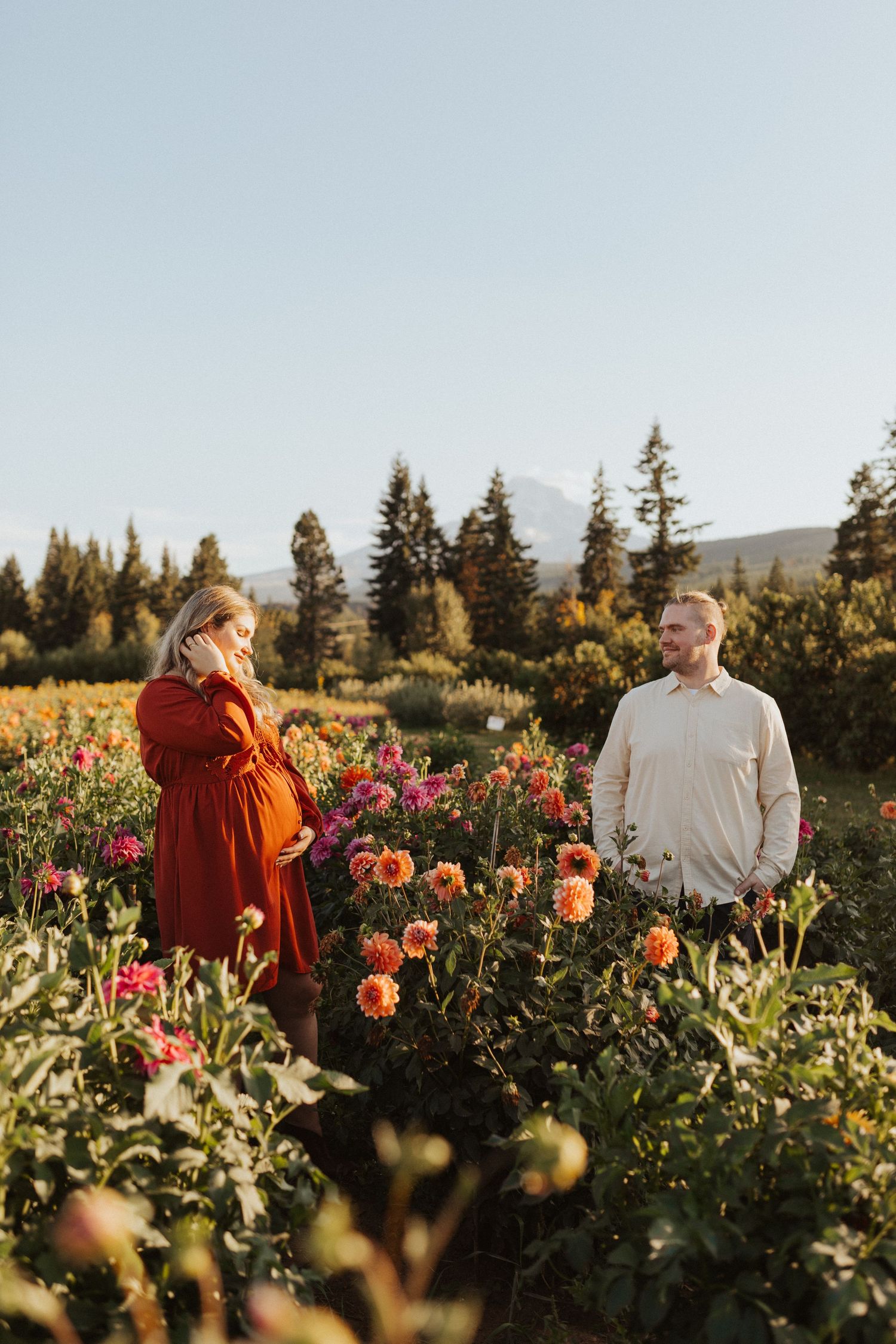 Expecting couple standing apart in a blooming dahlia field with evergreen trees and mountain backdrop.