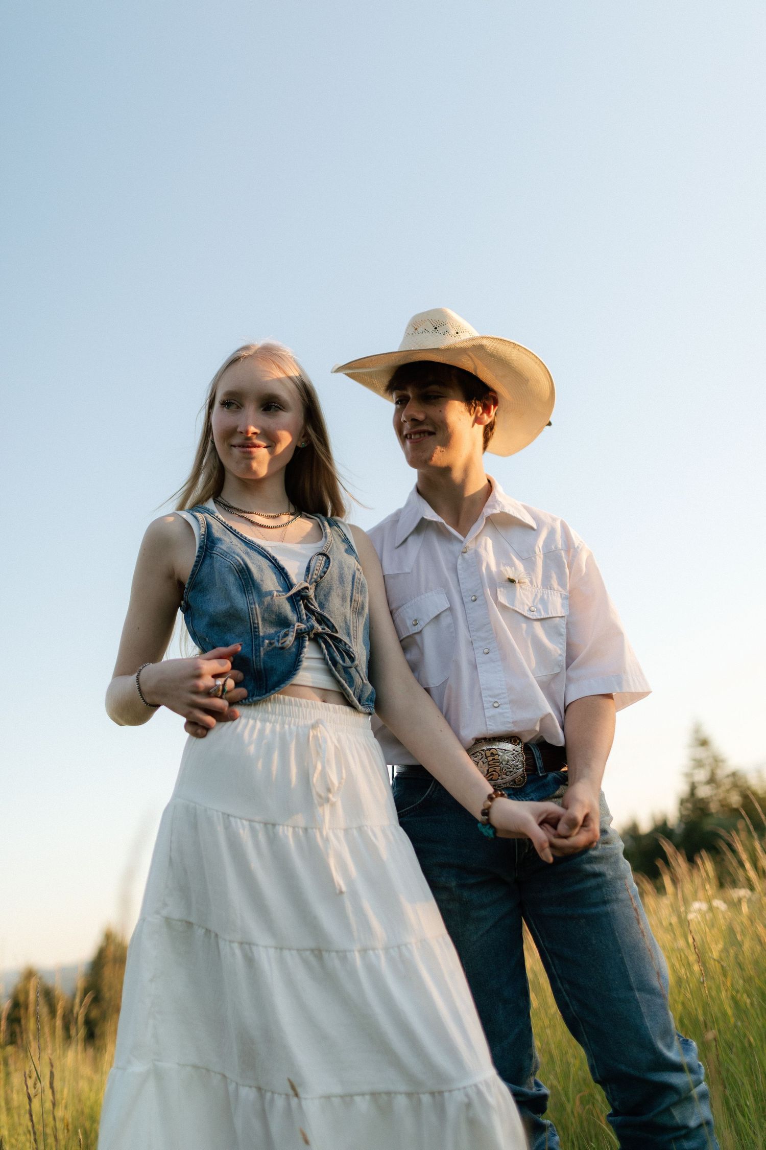 Young couple standing together in meadow at sunset, woman in white dress, man in cowboy hat smiling.