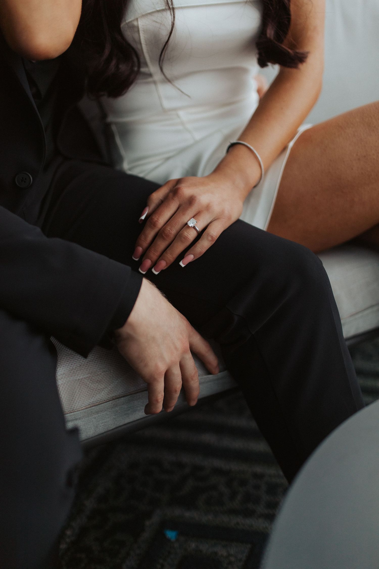 Close-up of couples hands showing diamond engagement ring, woman in white dress sitting beside man in black suit.