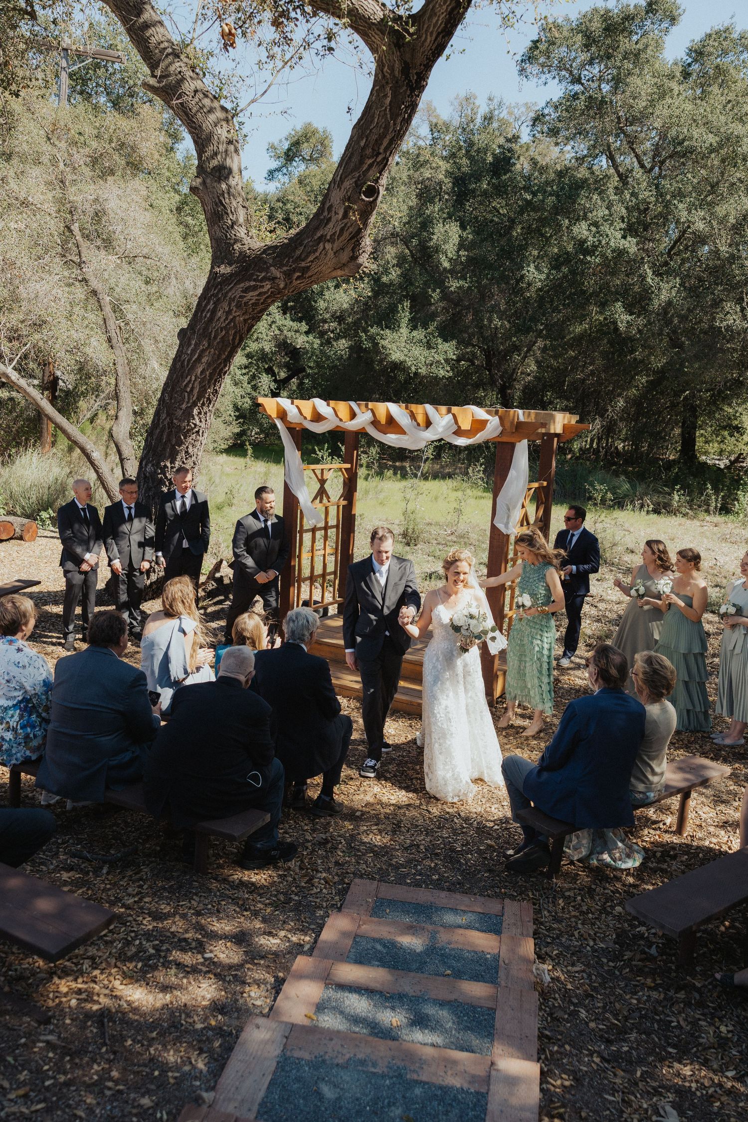 Outdoor wedding ceremony under wooden arch with guests seated, bride walking down the aisle.