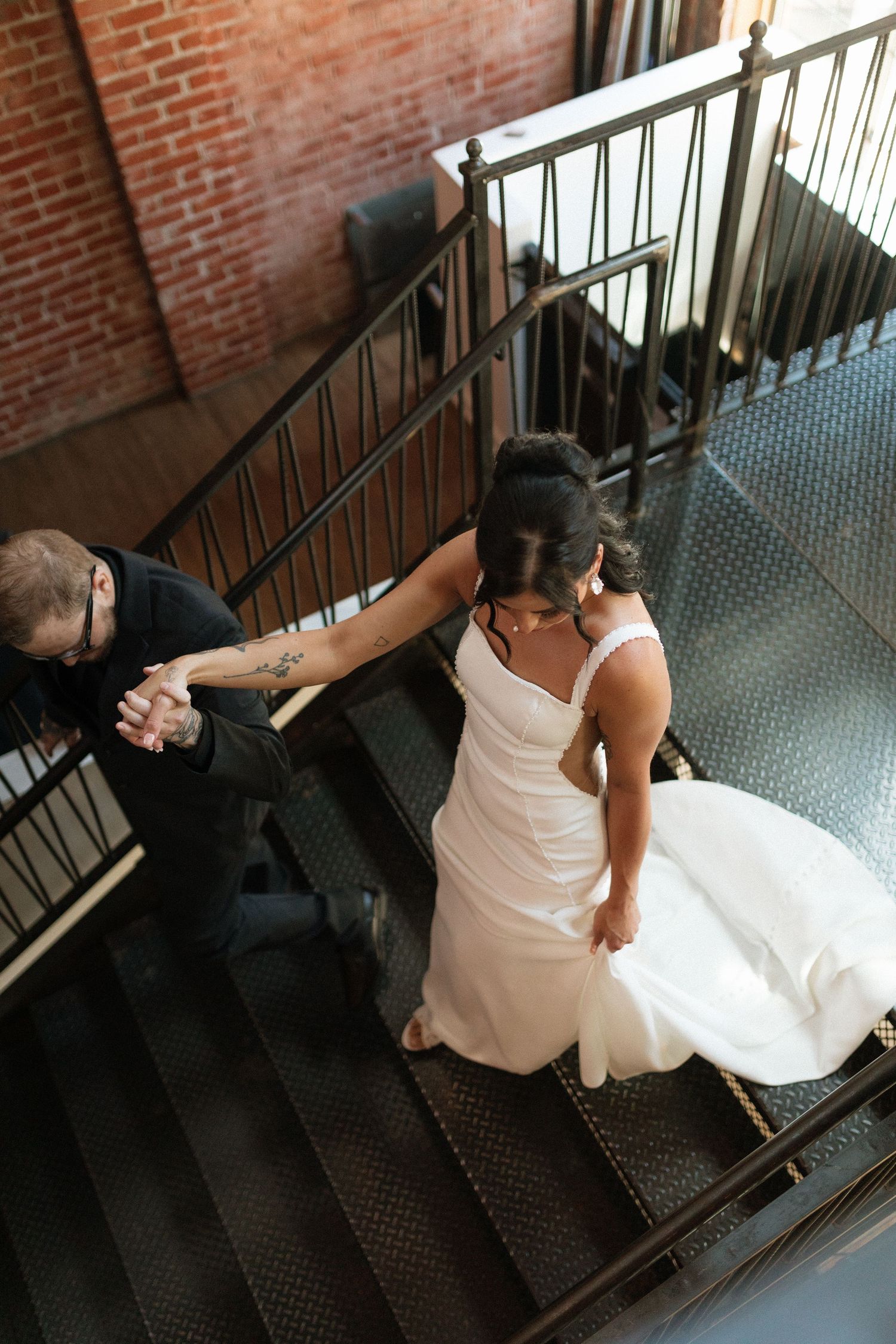 Aerial view of groom with tattoos helping bride in white gown descend iron staircase at venue.