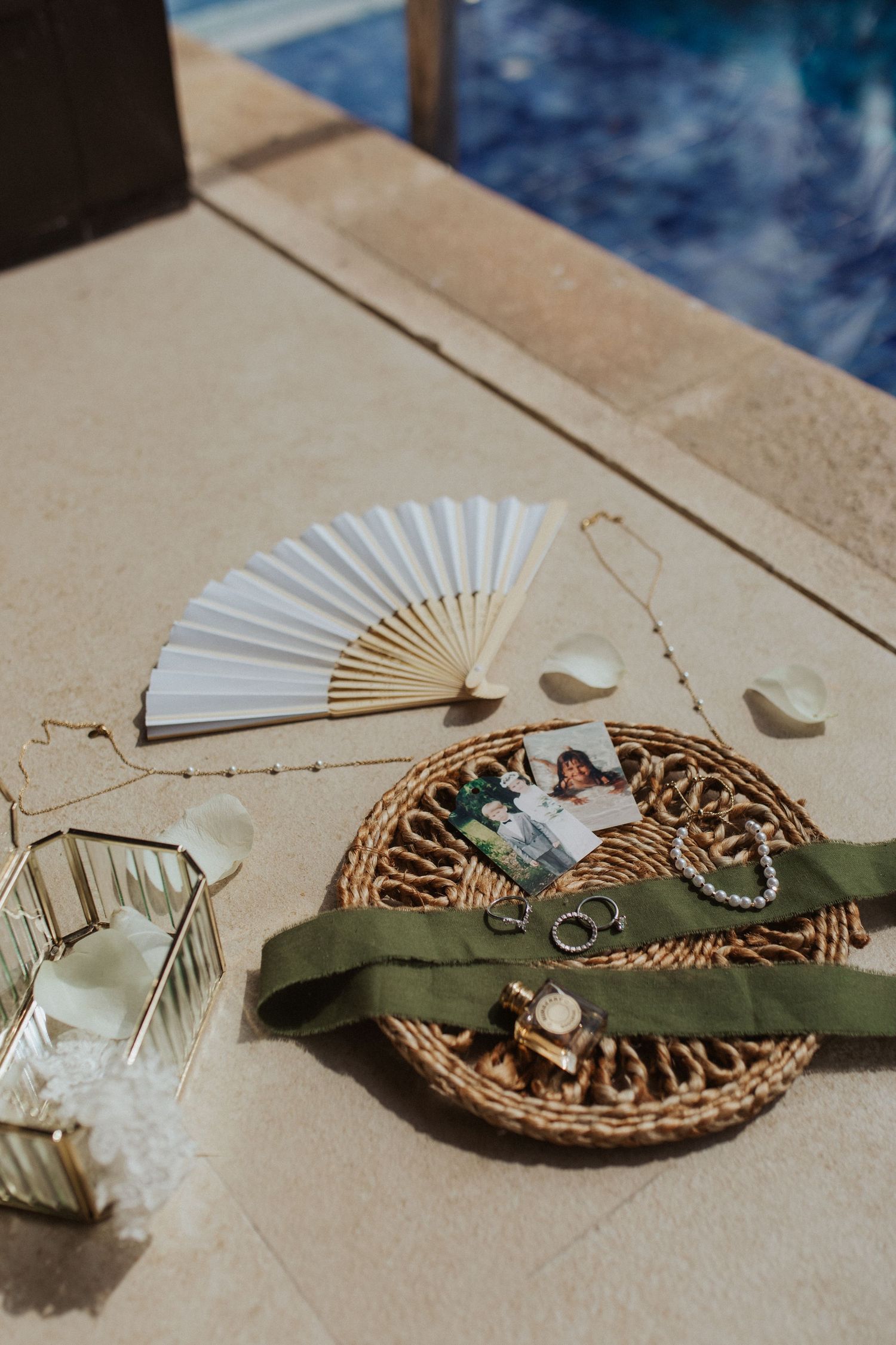 Wedding flat lay with white fan, jewelry, rings, green ribbon, and couple photo on wicker tray.