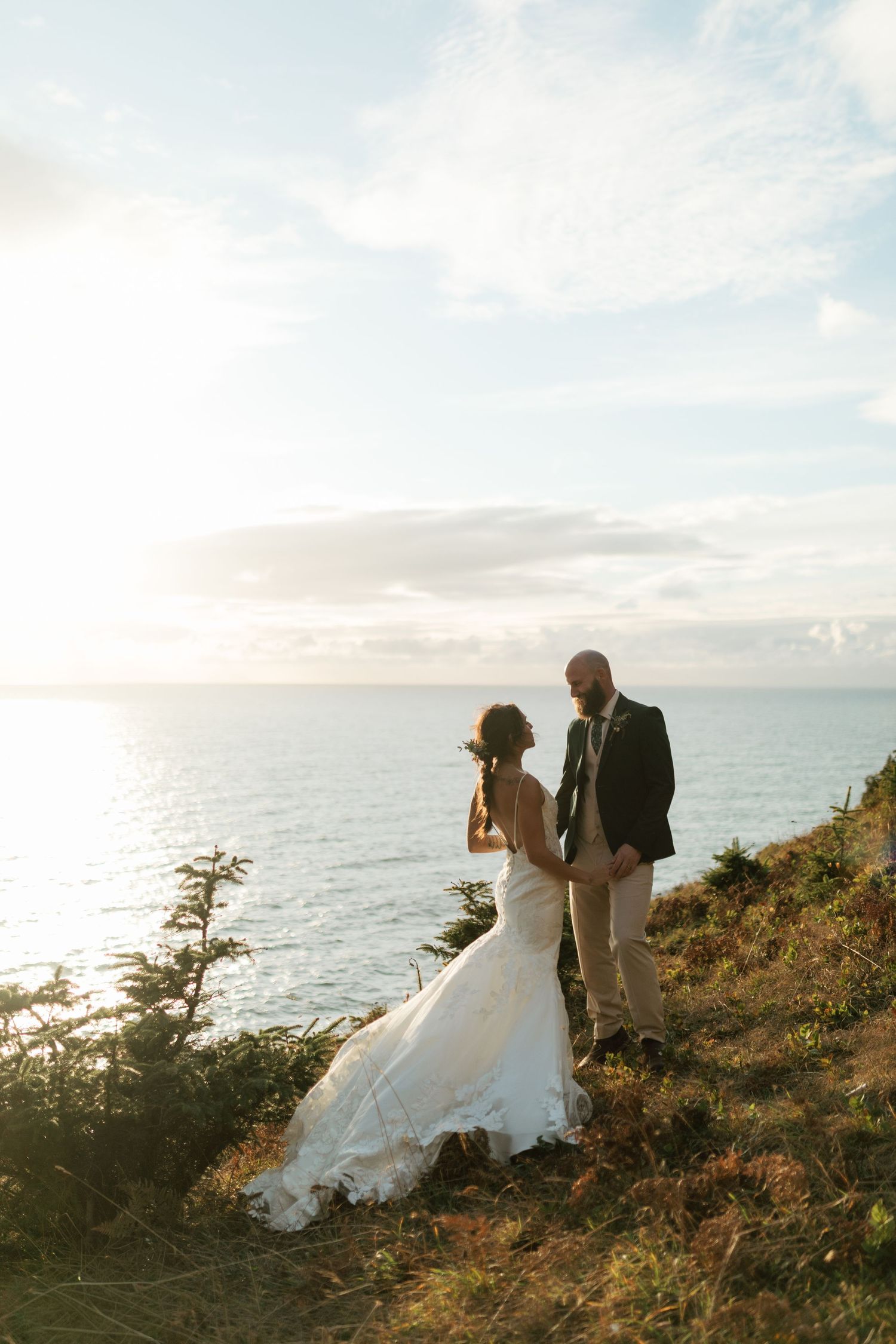 Bride and groom standing on a coastal cliff at sunset overlooking the ocean.