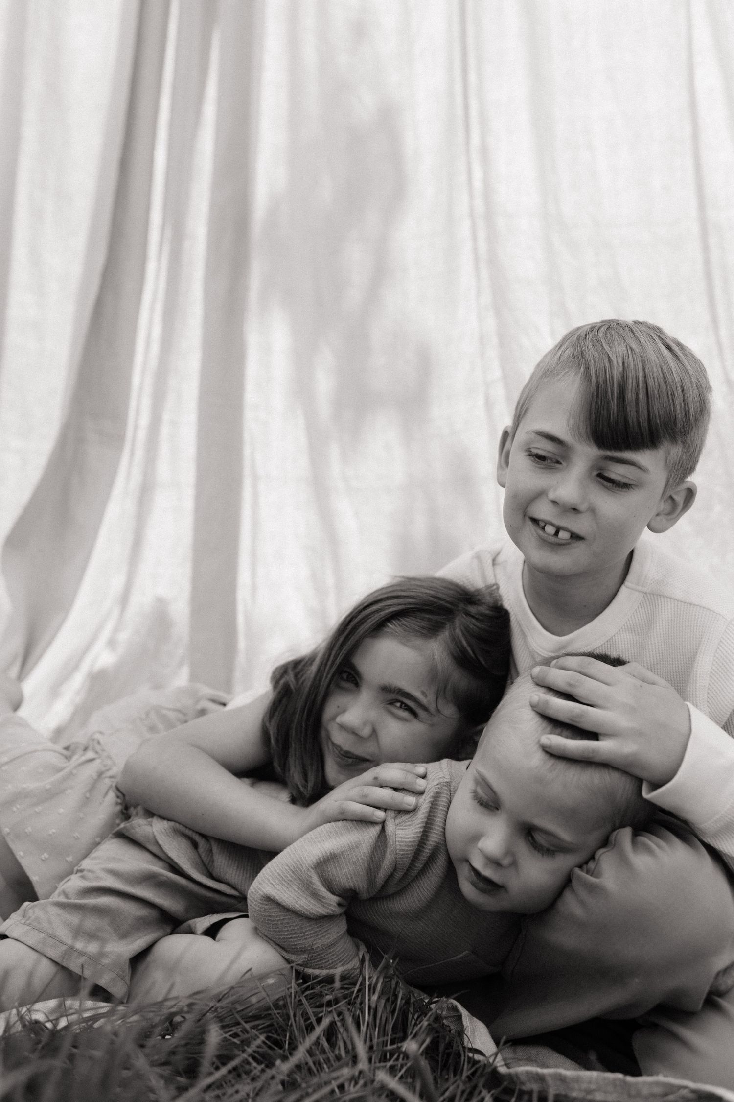 Black and white photo of three children laughing and cuddling together on grass outdoors.