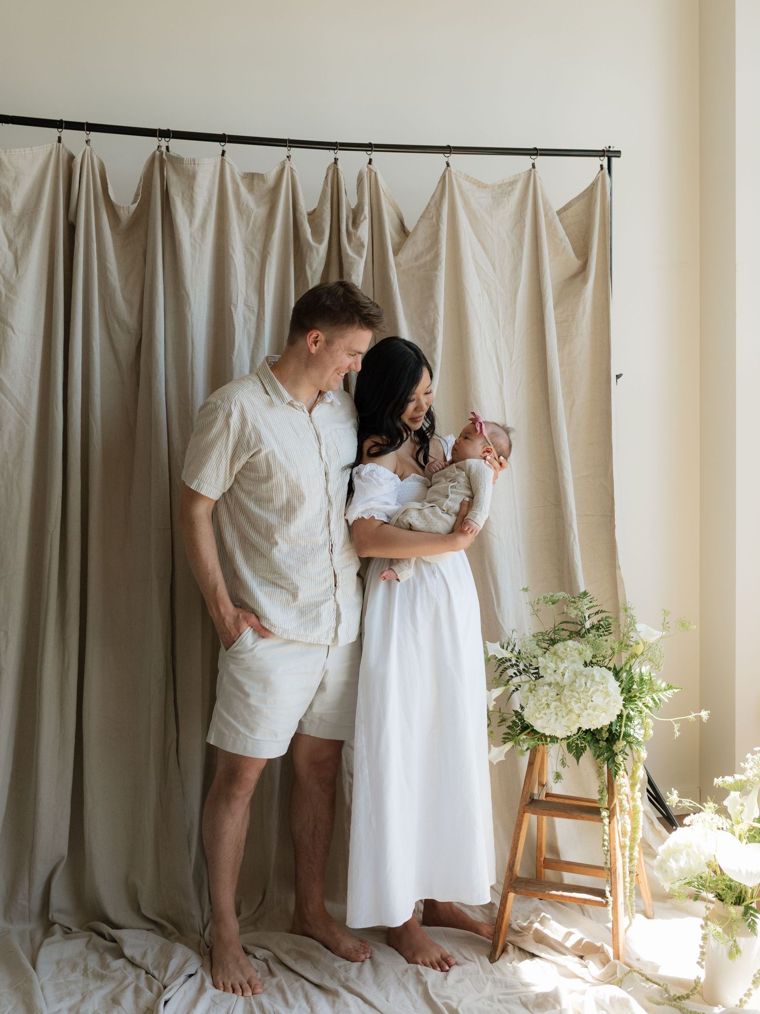A couple stands barefoot holding their newborn baby in a softly lit studio setting.