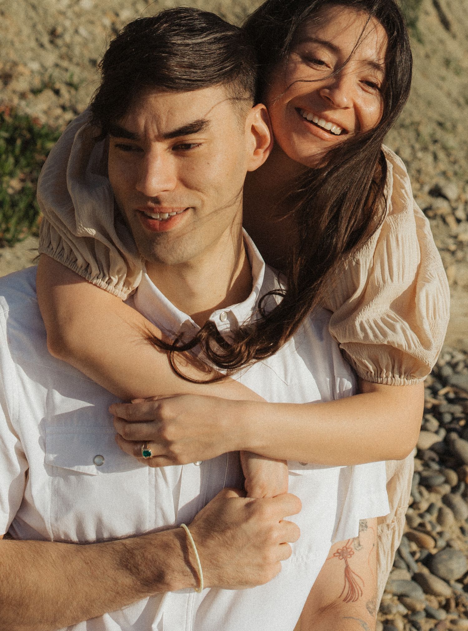 Laughing woman hugging man from behind outdoors, her hair blowing in the wind during a sunny beach session.