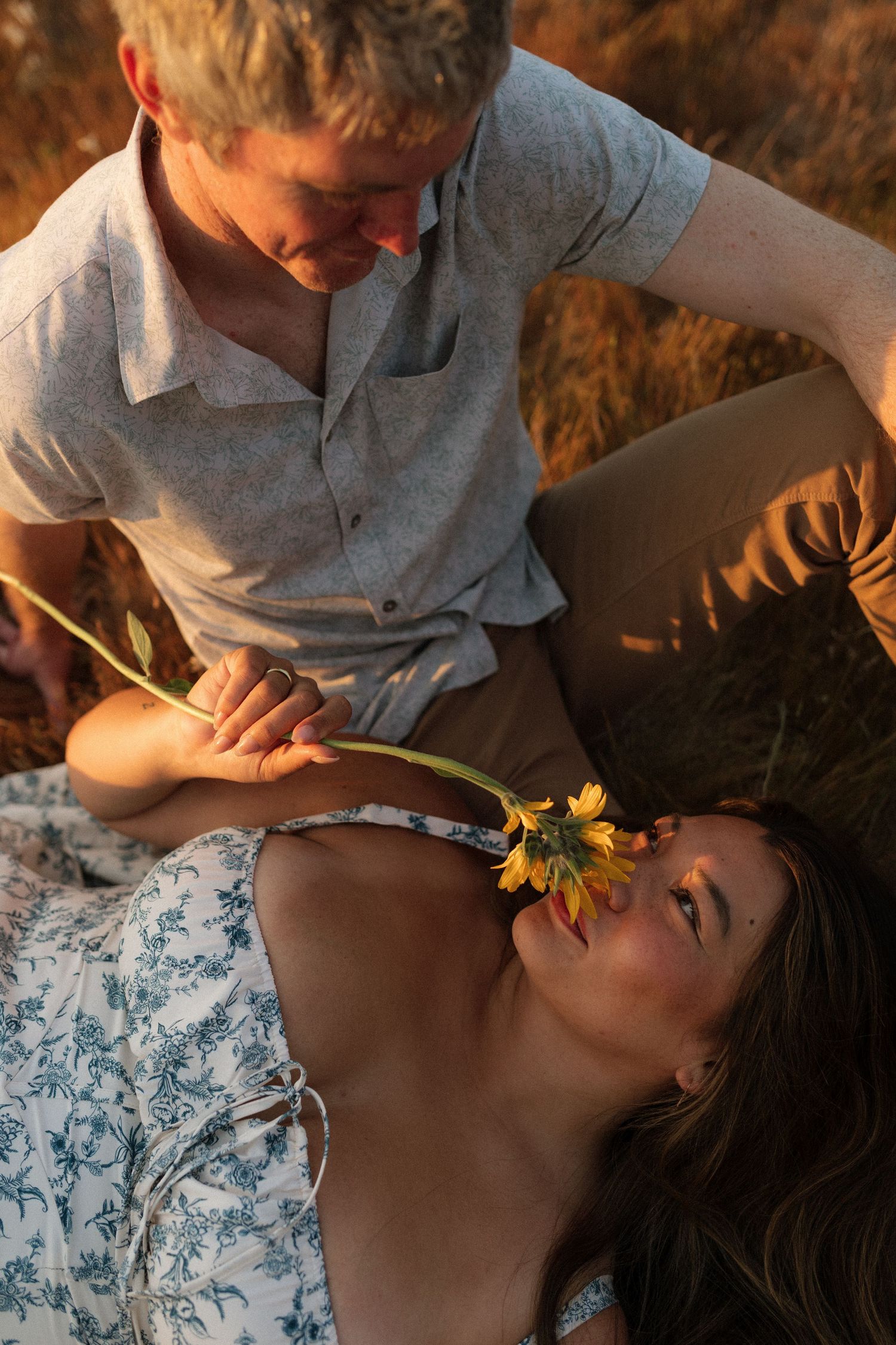 Man tenderly holding a sunflower over a woman lying in a golden field at sunset.