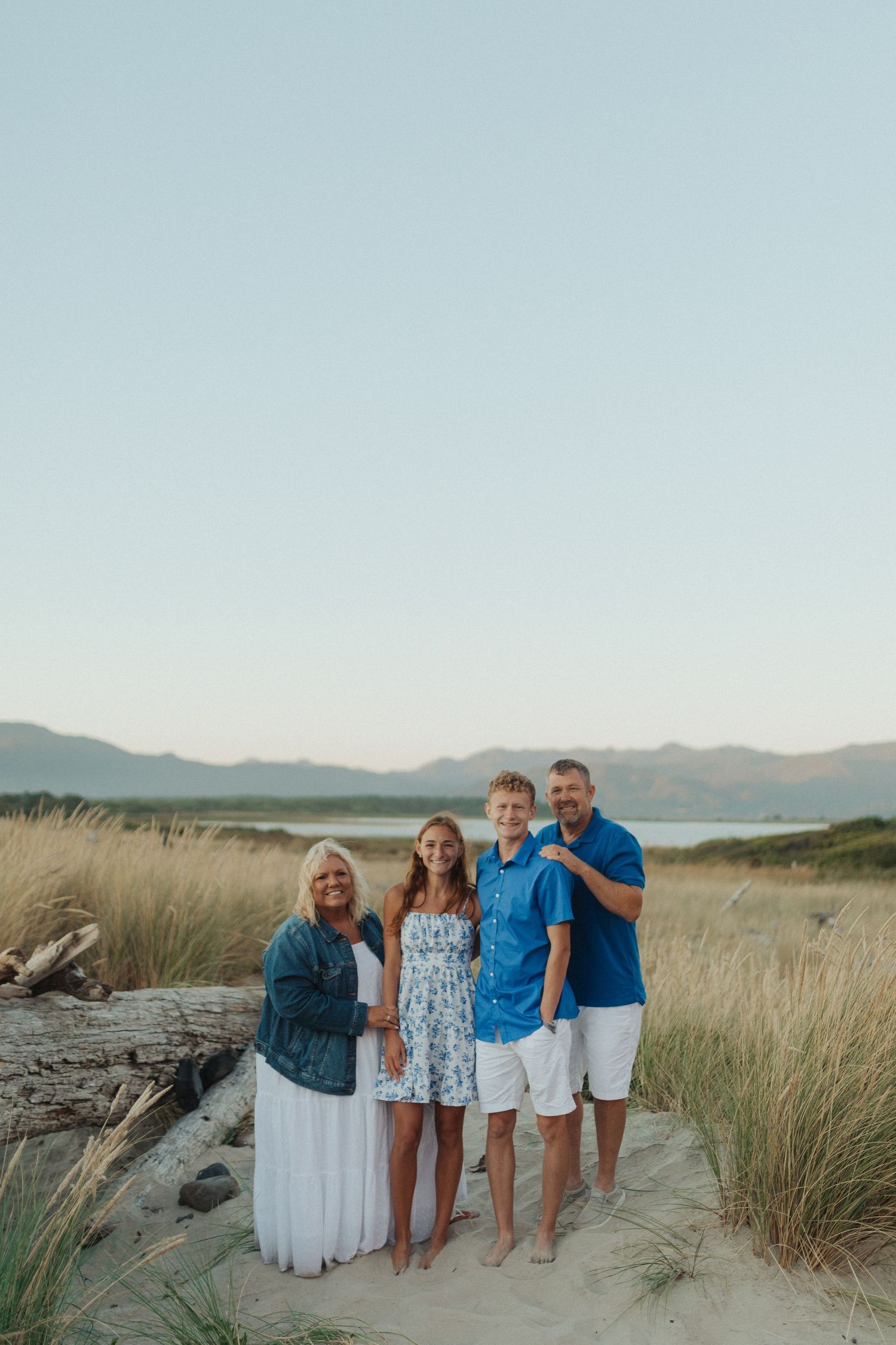 Family of four posing together on sandy beach with tall grass and mountain views at golden hour.