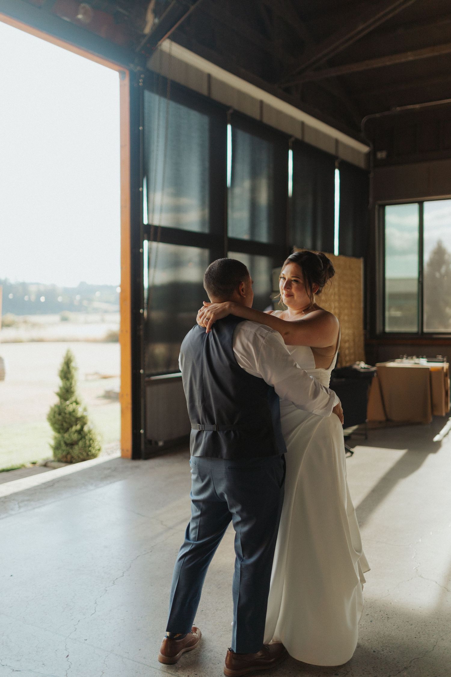 Bride and groom share first dance in a sunlit barn venue with large open doors revealing an outdoor view.