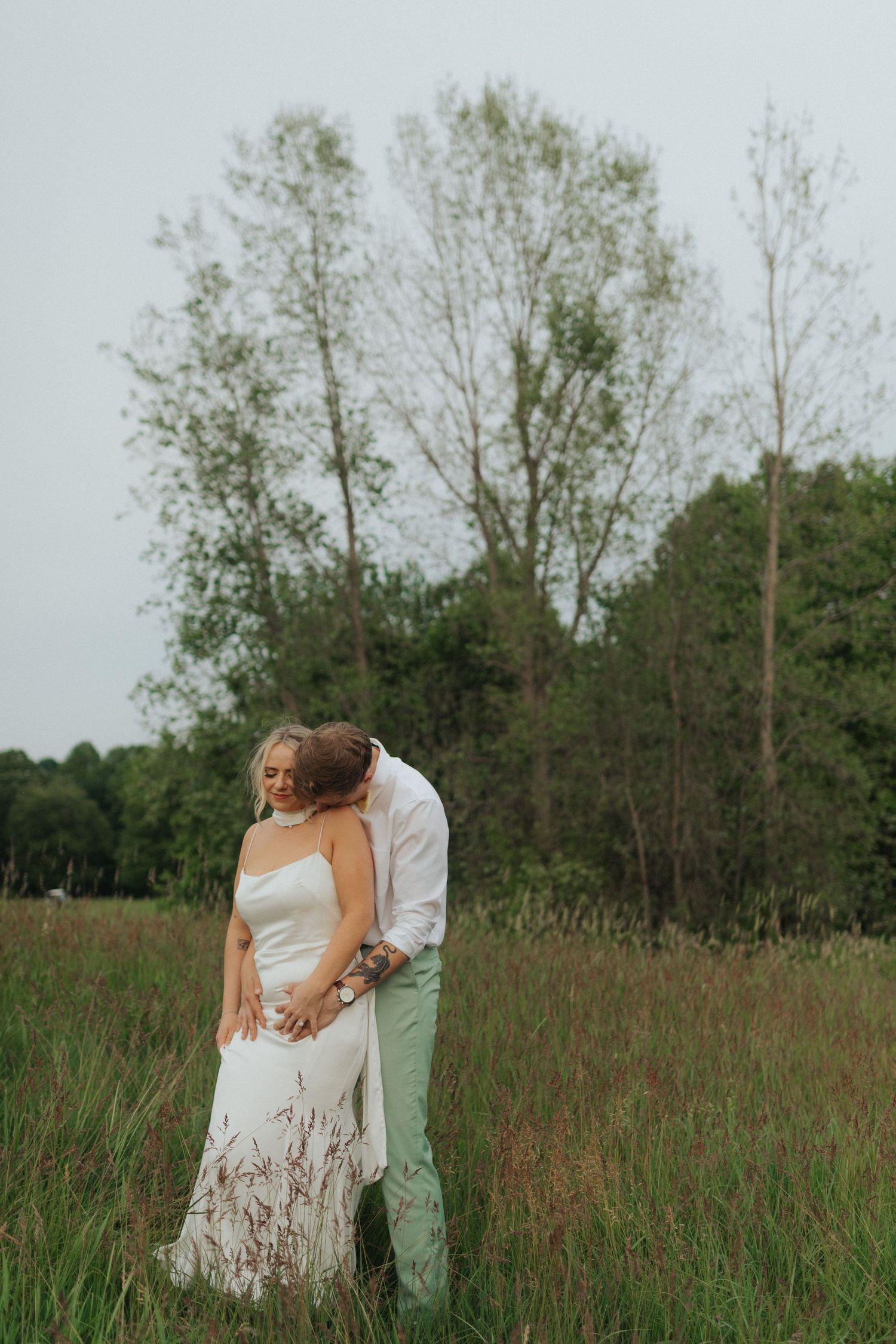 Bride and groom embrace in a lush green meadow, sharing a tender moment in wedding attire.