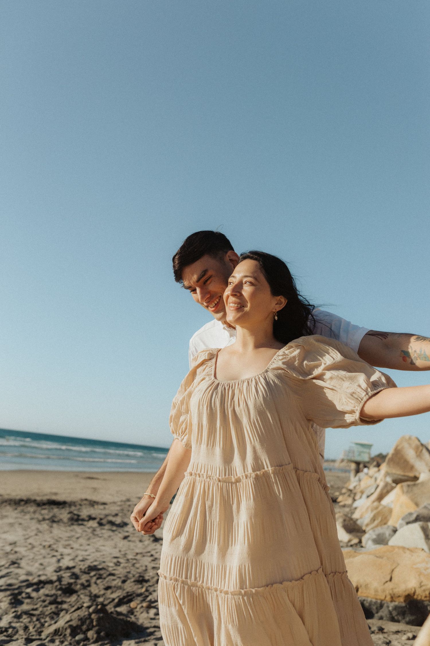 Man embracing woman from behind on a rocky beach, both smiling warmly under a clear blue sky.