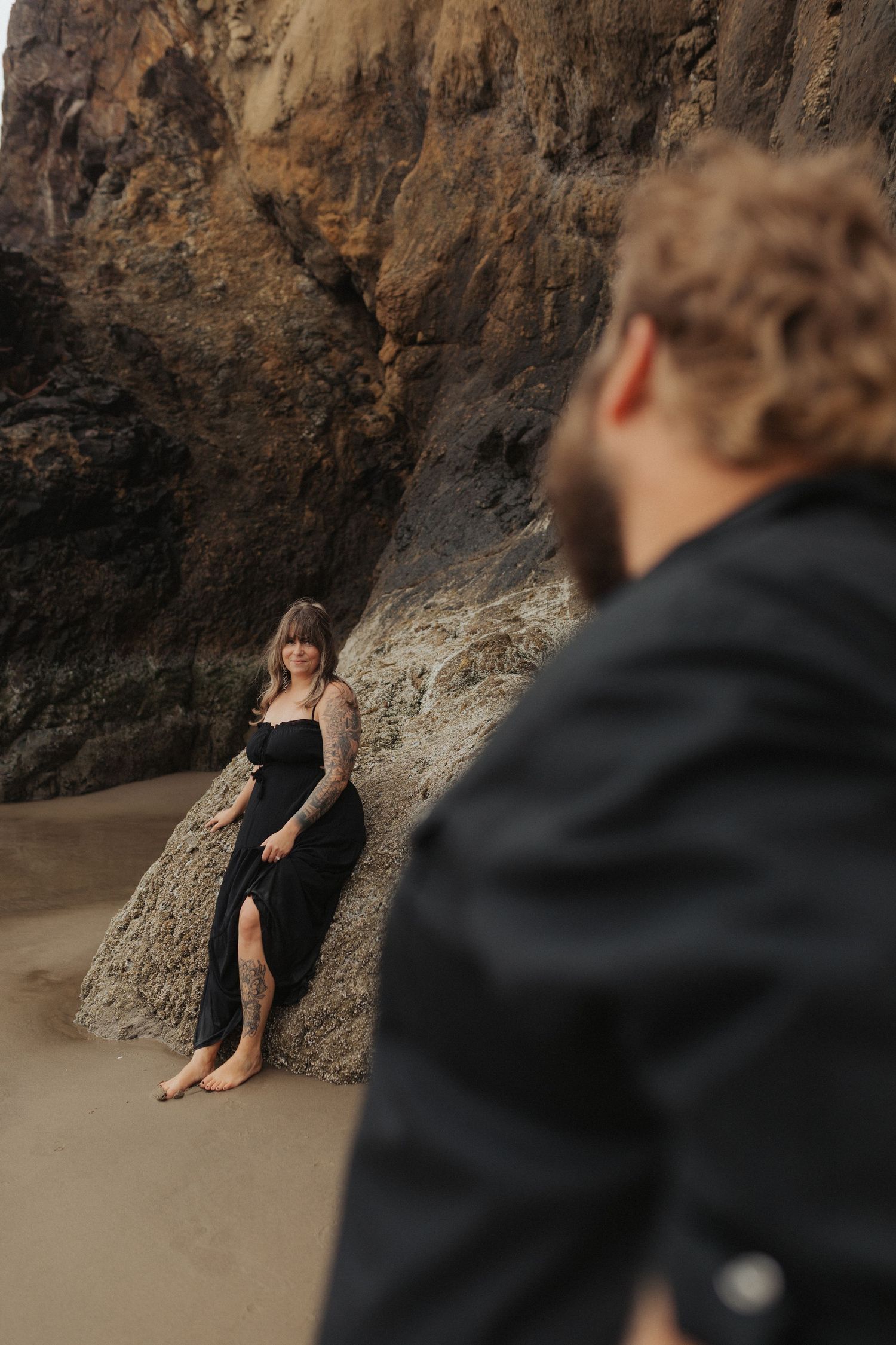 Tattooed woman in black dress leaning against beach rock formation, smiling at bearded partner.