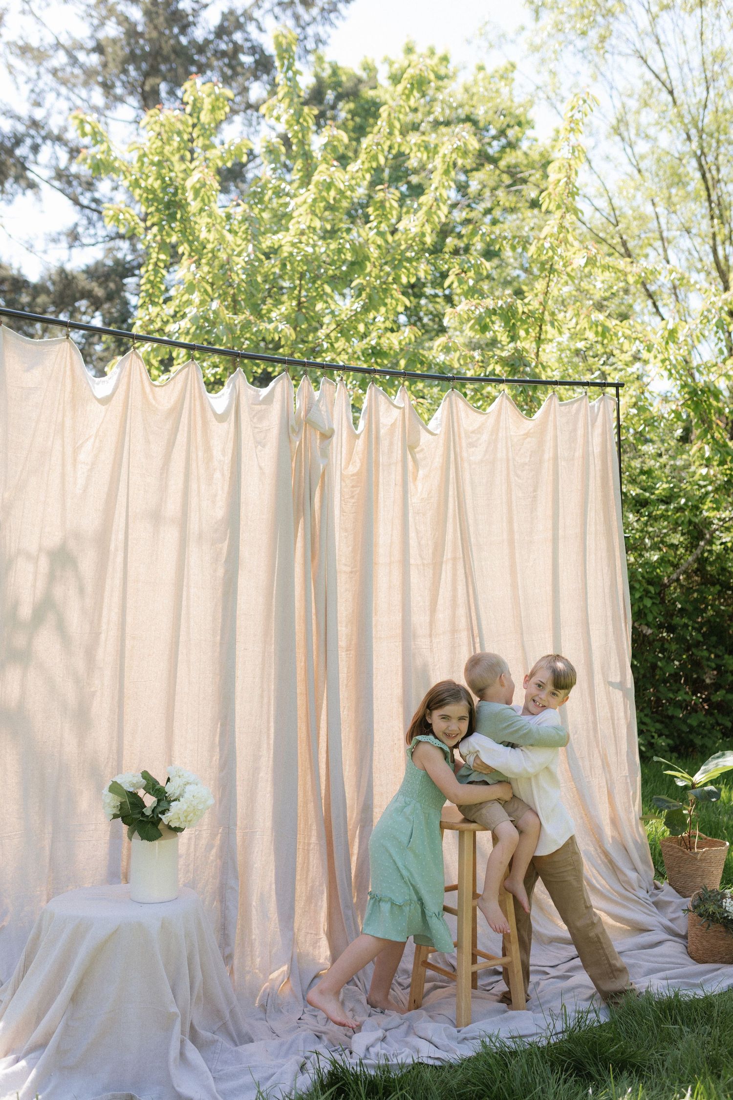 Two children embrace on a stool in front of a beige curtain backdrop in a sunlit garden setting.