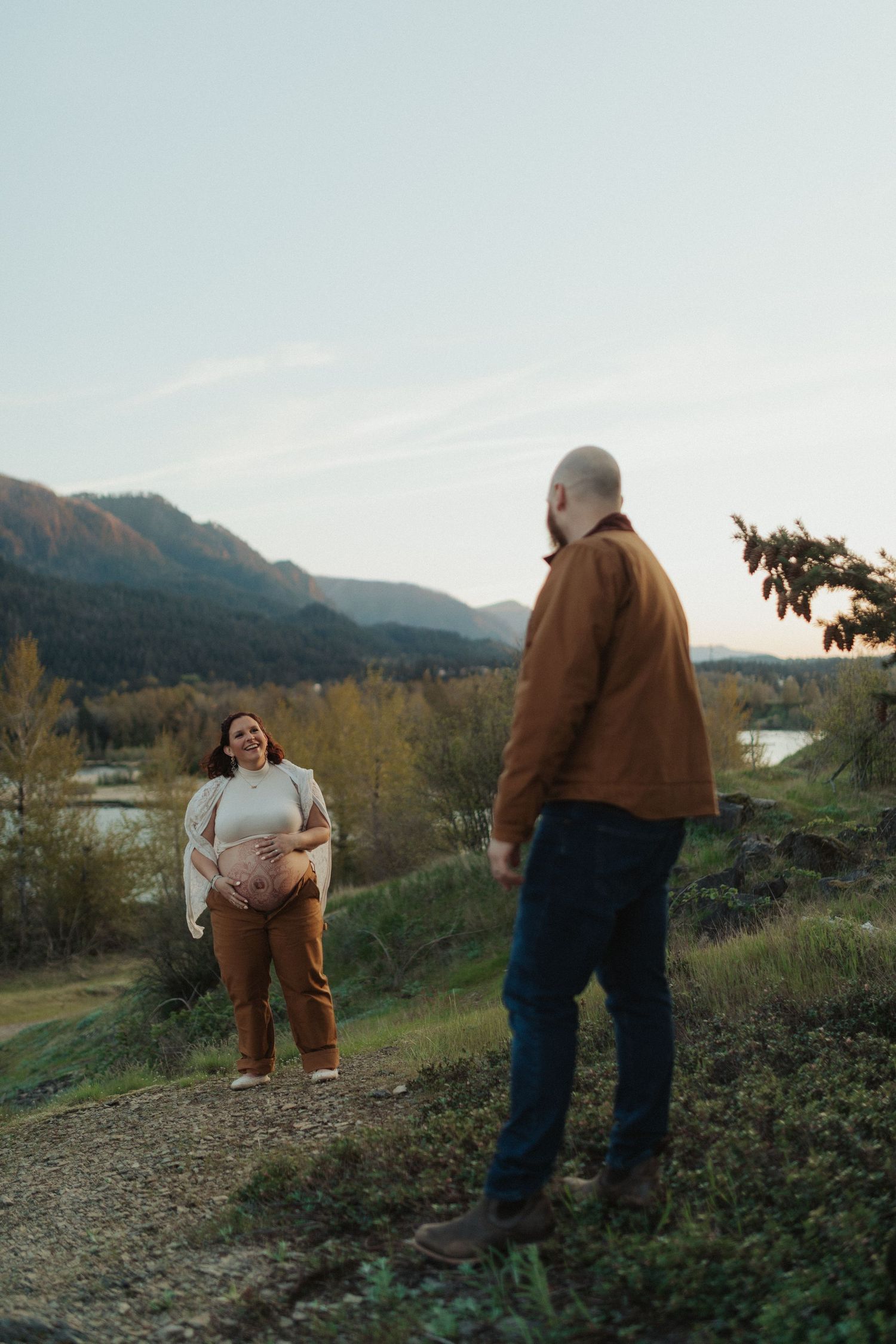 A pregnant woman cradles her baby bump while her partner stands facing her on a mountain overlook.
