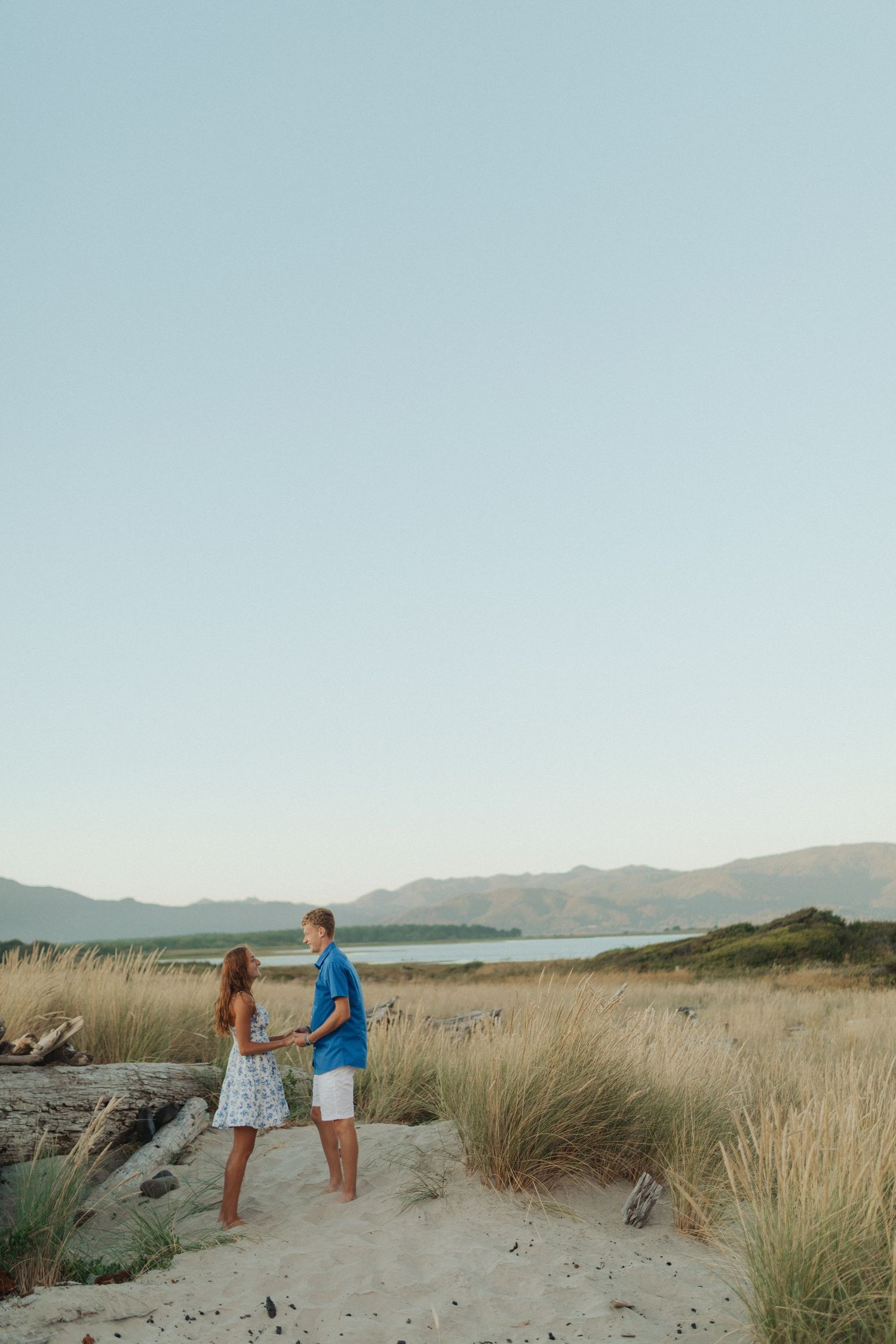 Young couple holding hands facing each other in sandy beach dunes with mountains in background.