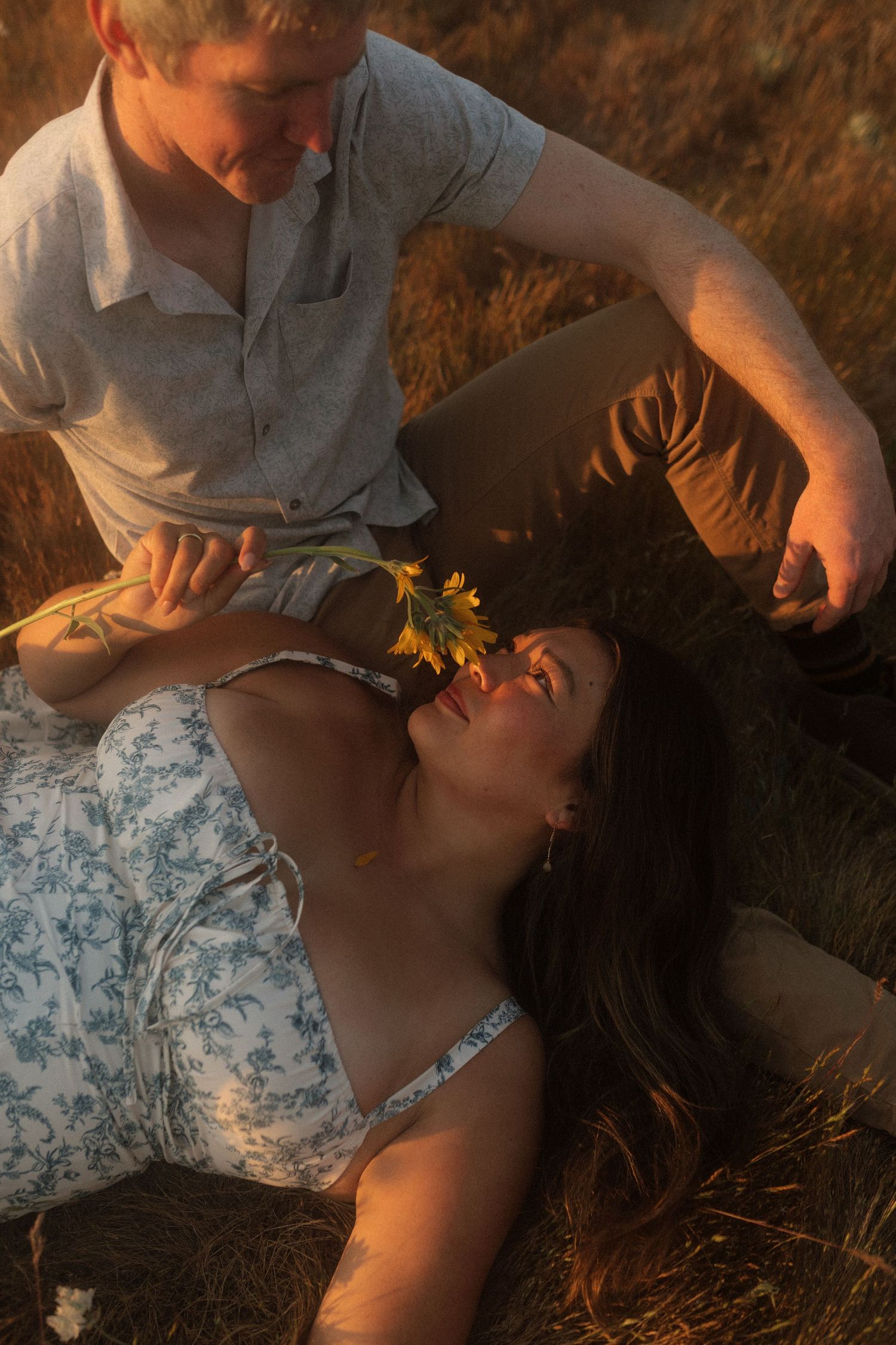 Romantic couple in a sunlit field, man offering wildflowers to woman lying in golden grass.