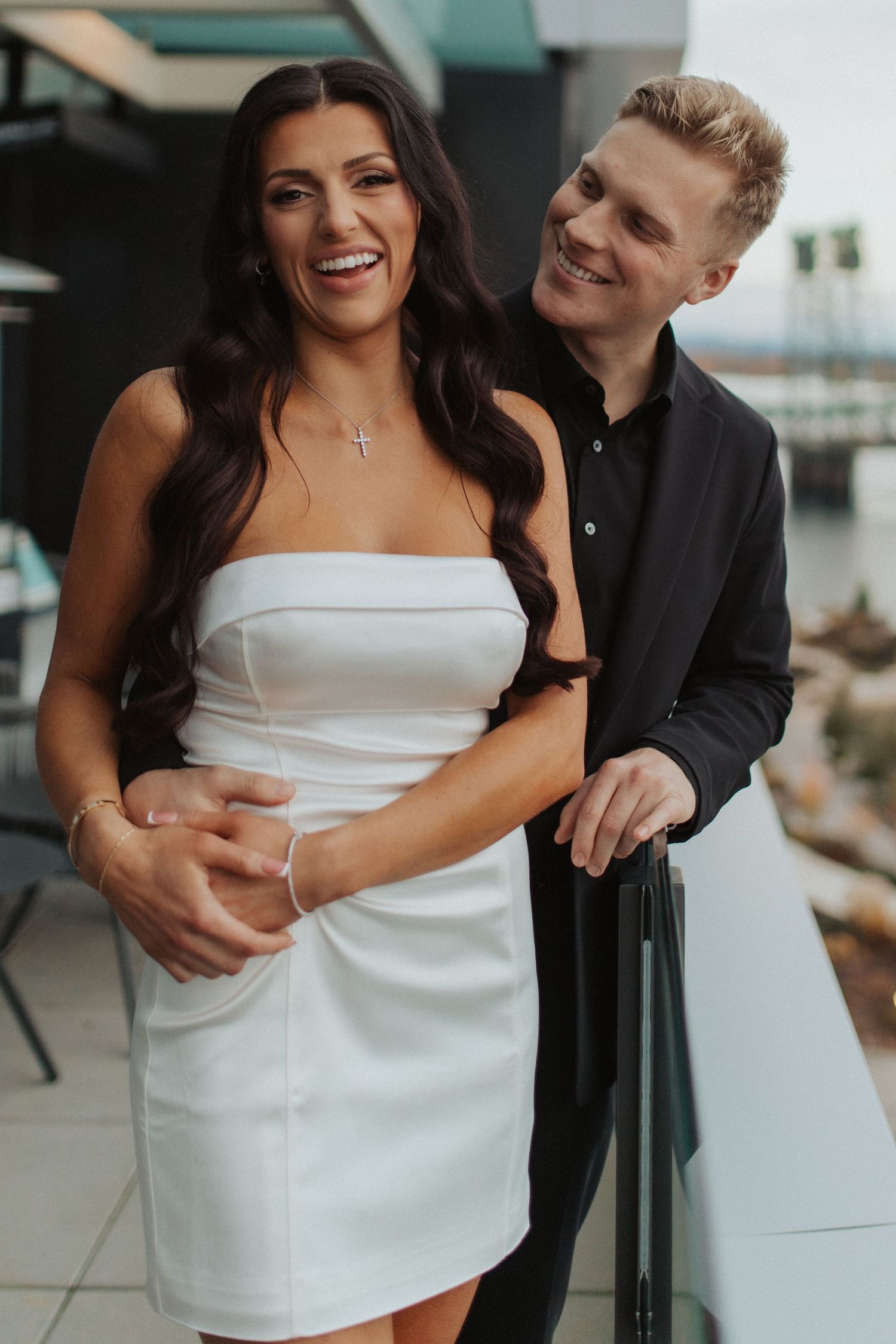 Happy couple embracing on outdoor terrace, woman in white strapless dress laughing with man in black jacket.